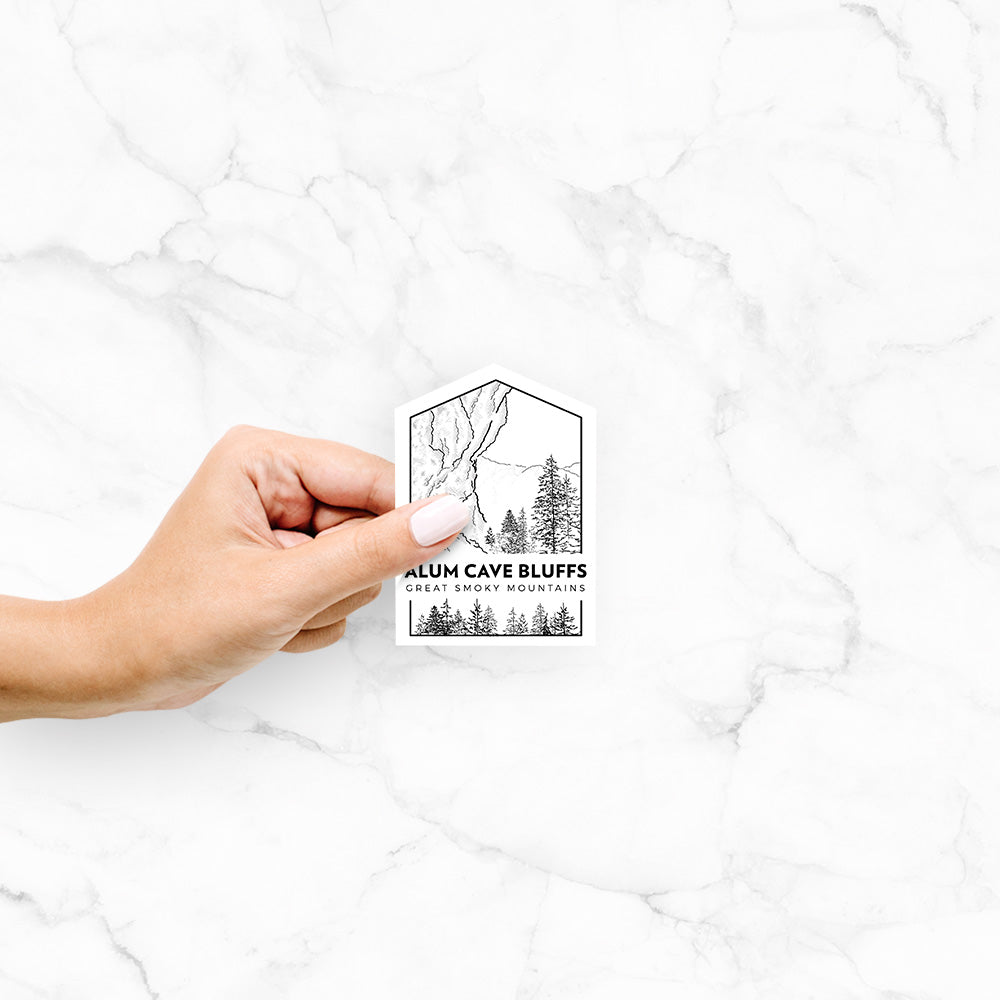 Hand holding a 4 inch vinyl decal of a black line art badge of Alum Cave Bluffs with pine trees, with the words Alum Cave Bluffs, Great Smoky Mountains.