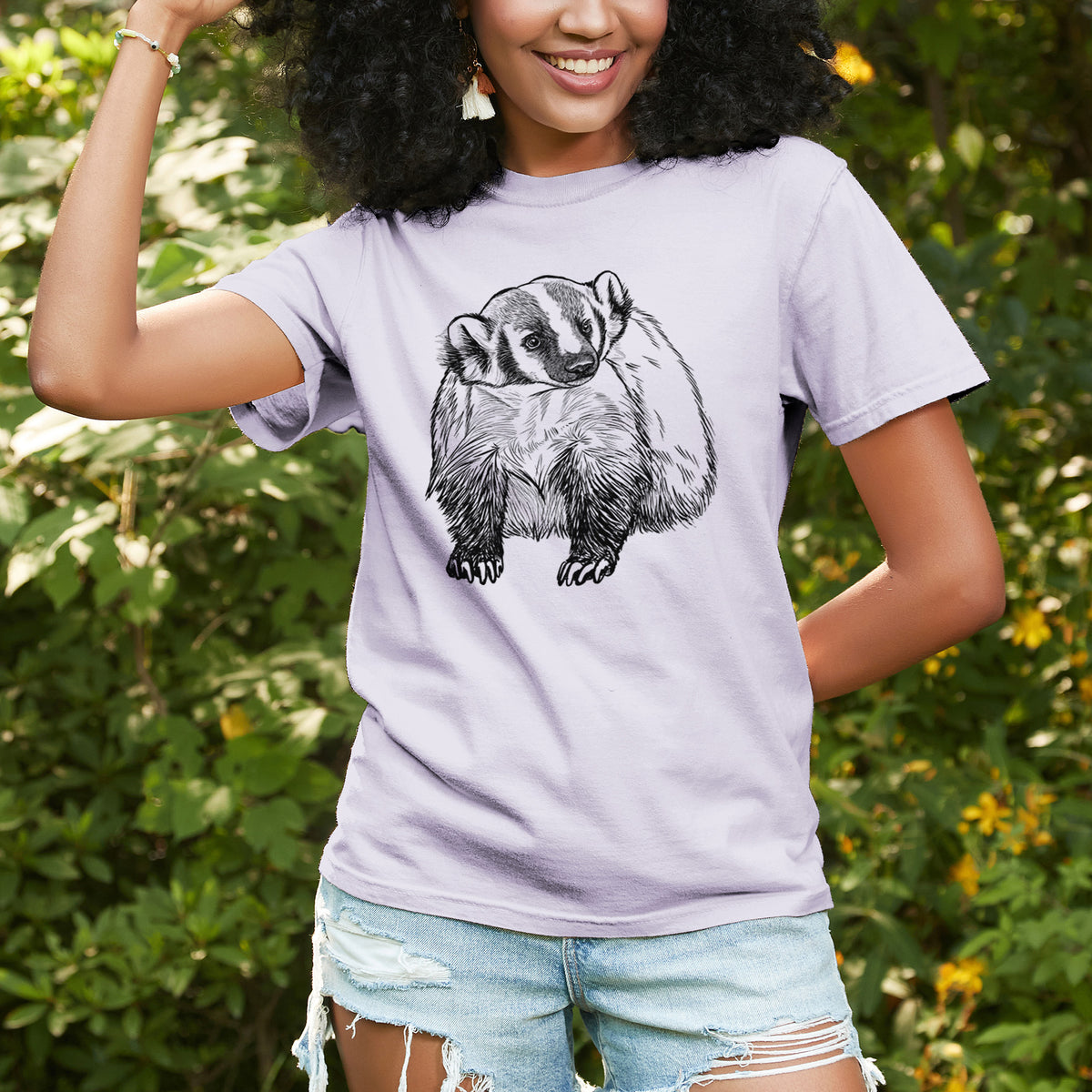 Woman wearing a Comfort Colors t-shirt printed with a detailed black ink illustration of an American badger facing forward, outdoors.