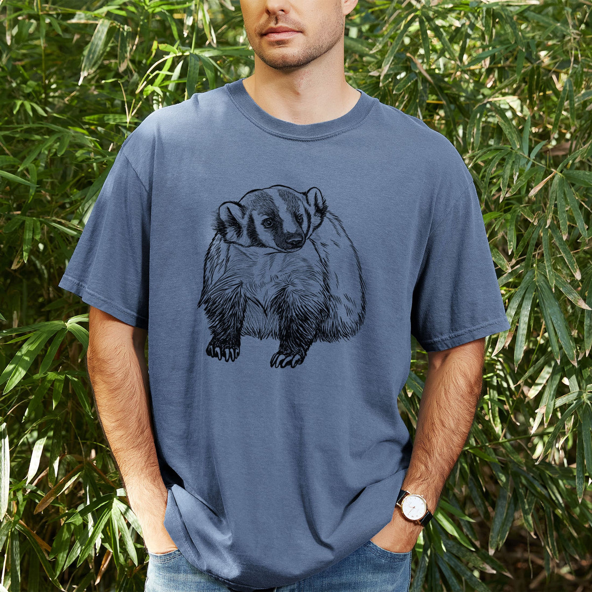 Man wearing a Comfort Colors t-shirt printed with a detailed black ink illustration of an American badger facing forward, outdoors.