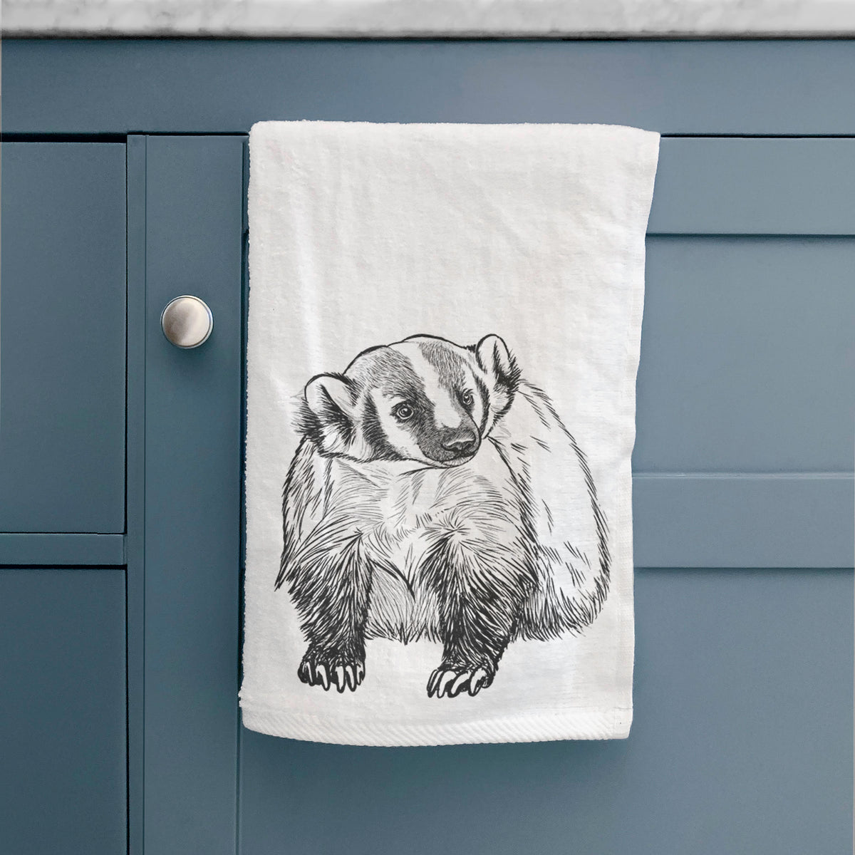 White hand towel printed with a detailed black ink illustration of an American badger facing forward, hanging on a cabinet door.