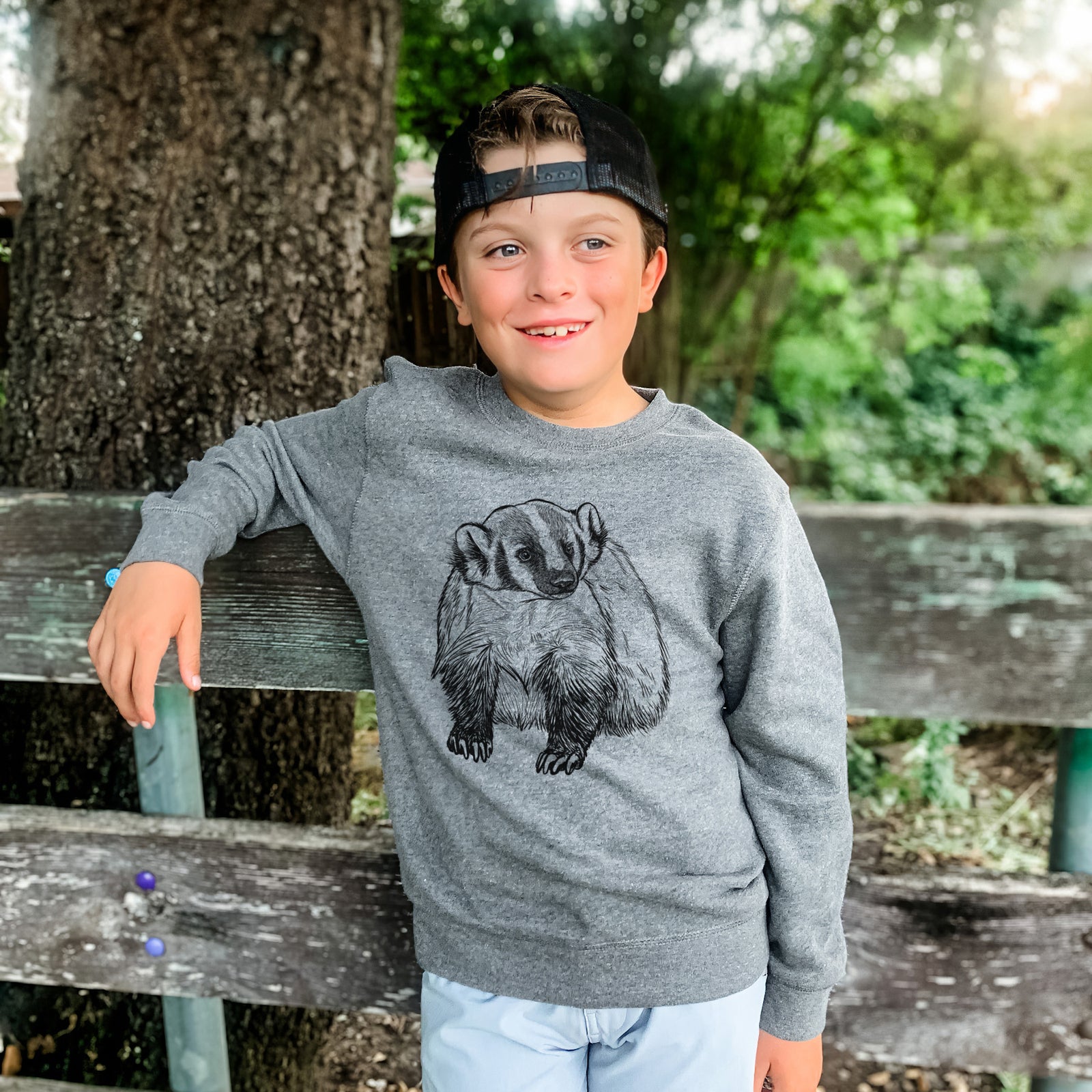 Boy wearing a youth crewneck sweatshirt printed with a detailed black ink illustration of an American badger facing forward, outdoors.