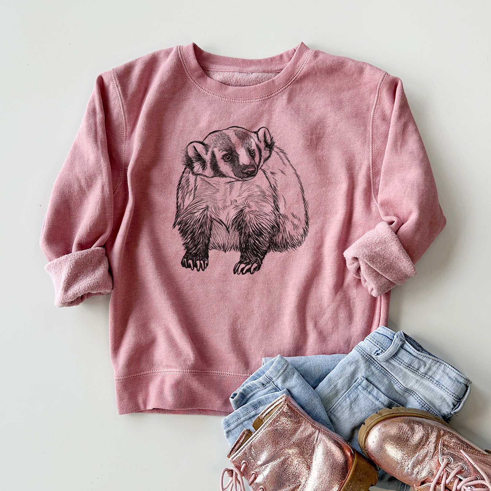 Boy wearing a youth crewneck sweatshirt printed with a detailed black ink illustration of an American badger facing forward, outdoors.