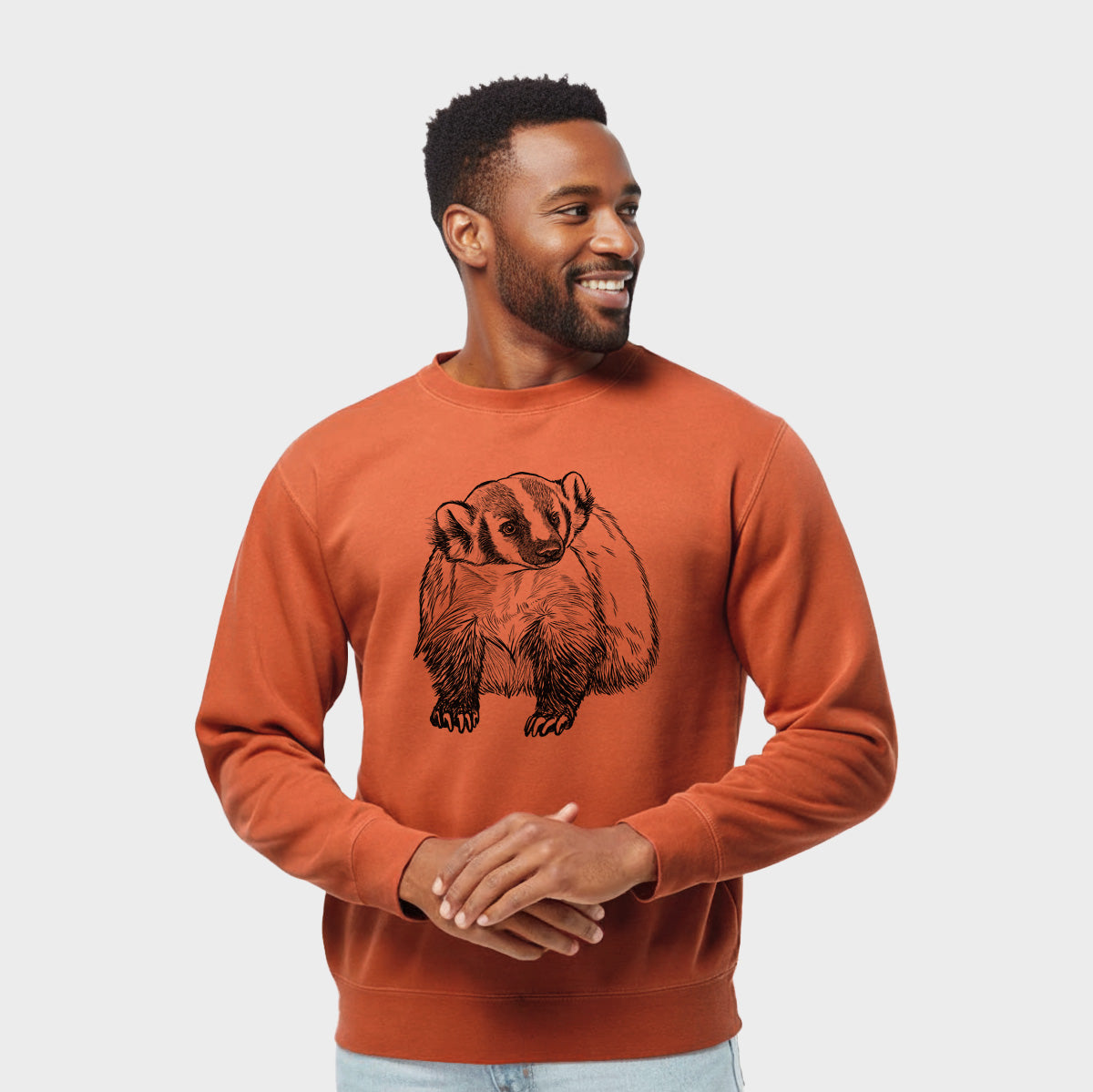 Model wearing an amber orange pigment dyed crewneck sweatshirt printed with a detailed black ink illustration of an American badger facing forward.