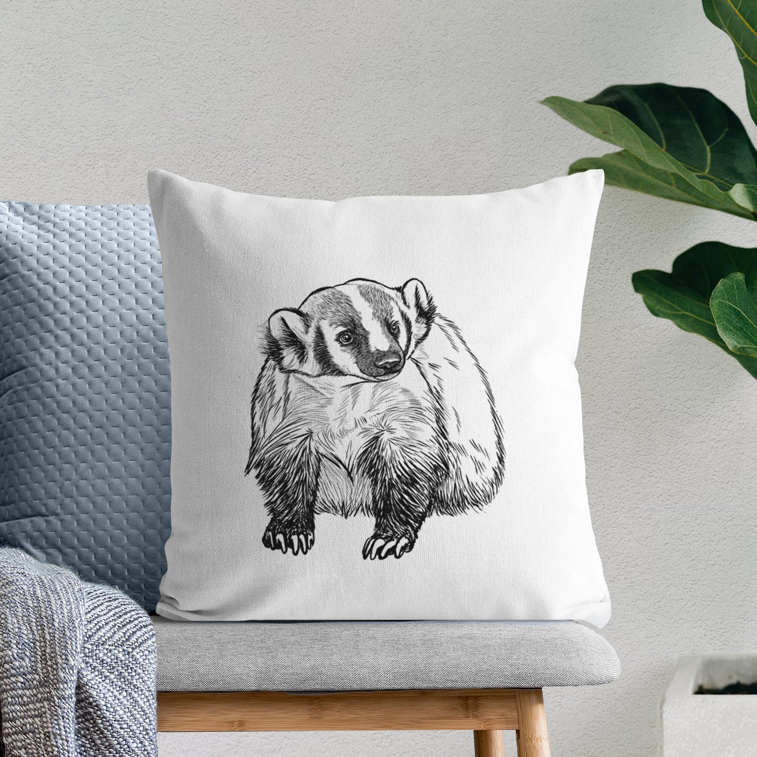 White pillow cover printed with a detailed black ink illustration of an American badger facing forward, on a bench beside plants.