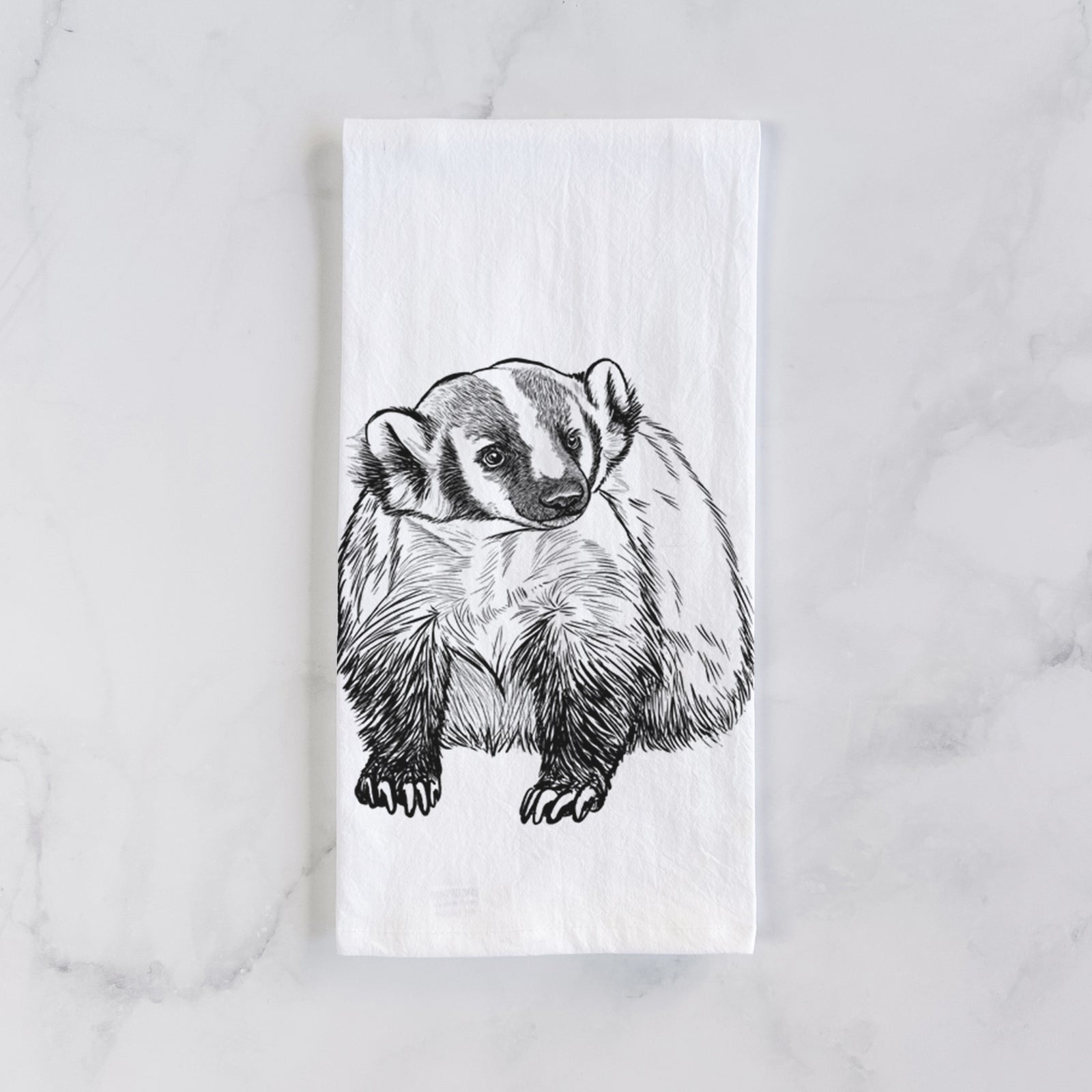 White cotton tea towel printed with a detailed black ink illustration of an American badger facing forward, on a marble counter beside a round wooden cutting board and greenery.
