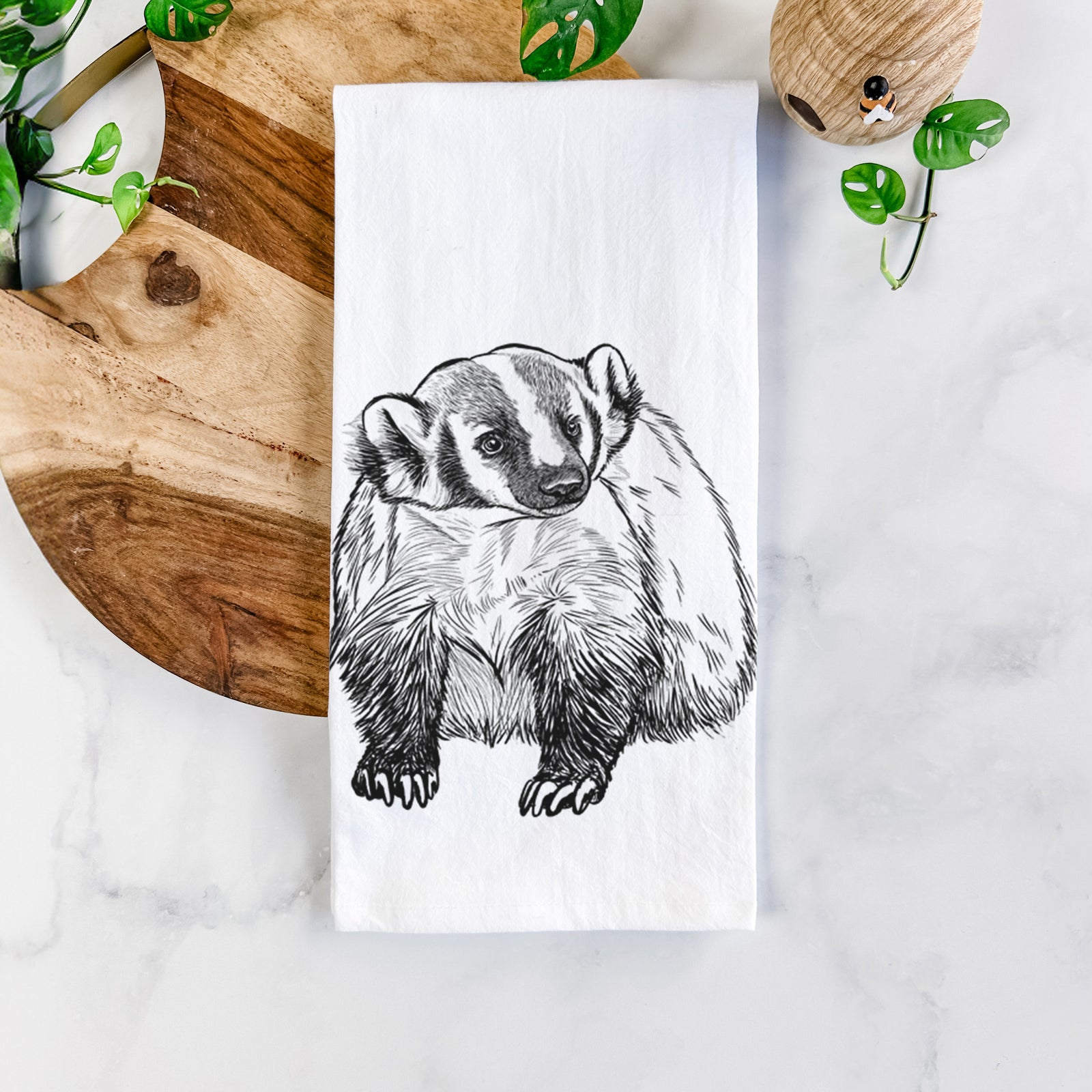 White cotton tea towel printed with a detailed black ink illustration of an American badger facing forward, on a marble counter beside a round wooden cutting board and greenery.