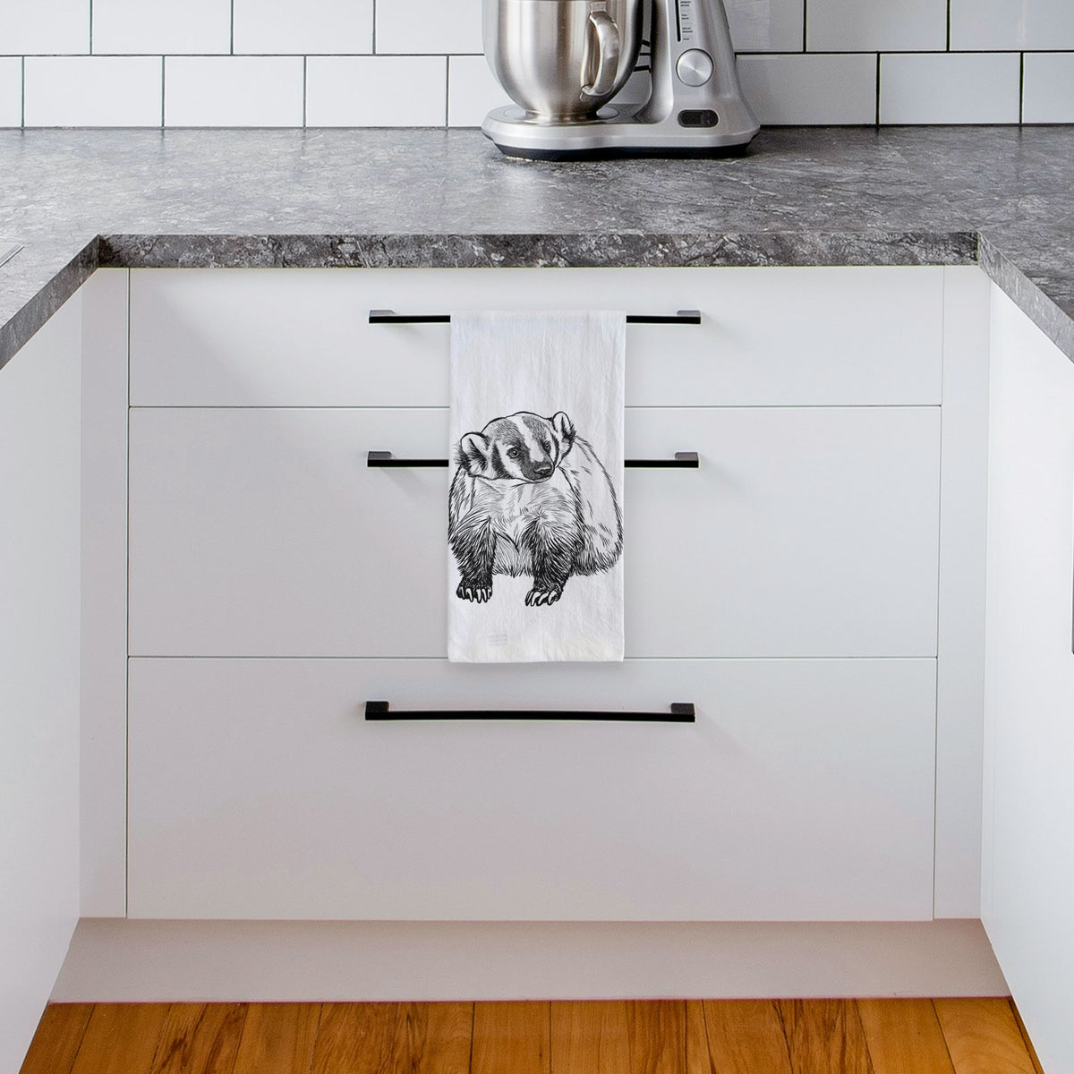 White tea towel printed with a detailed black ink illustration of an American badger facing forward, hanging from a drawer handle in a bright white kitchen.