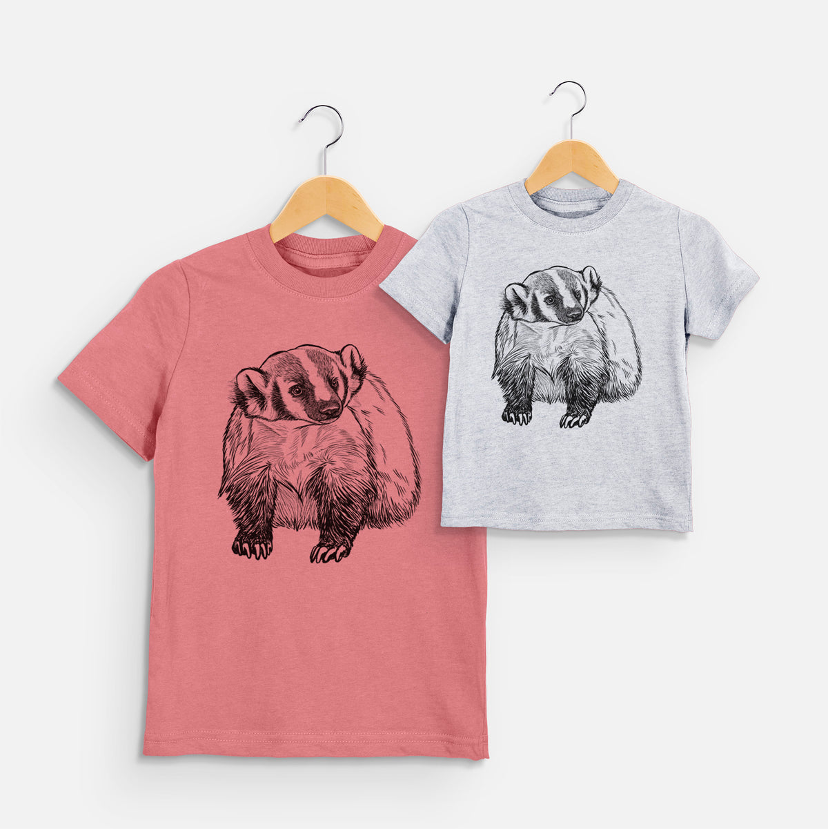 Two youth t-shirts in different colors on hangers, printed with a detailed black ink illustration of an American badger facing forward.