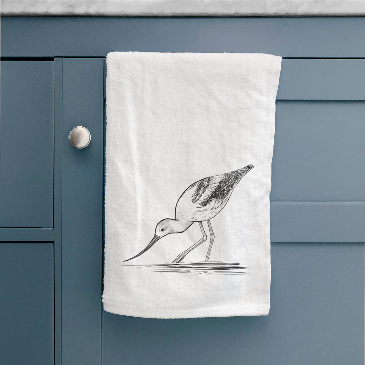 White hand towel printed with a detailed black ink sketch of an American avocet wading in shallow water, hanging on a cabinet door.