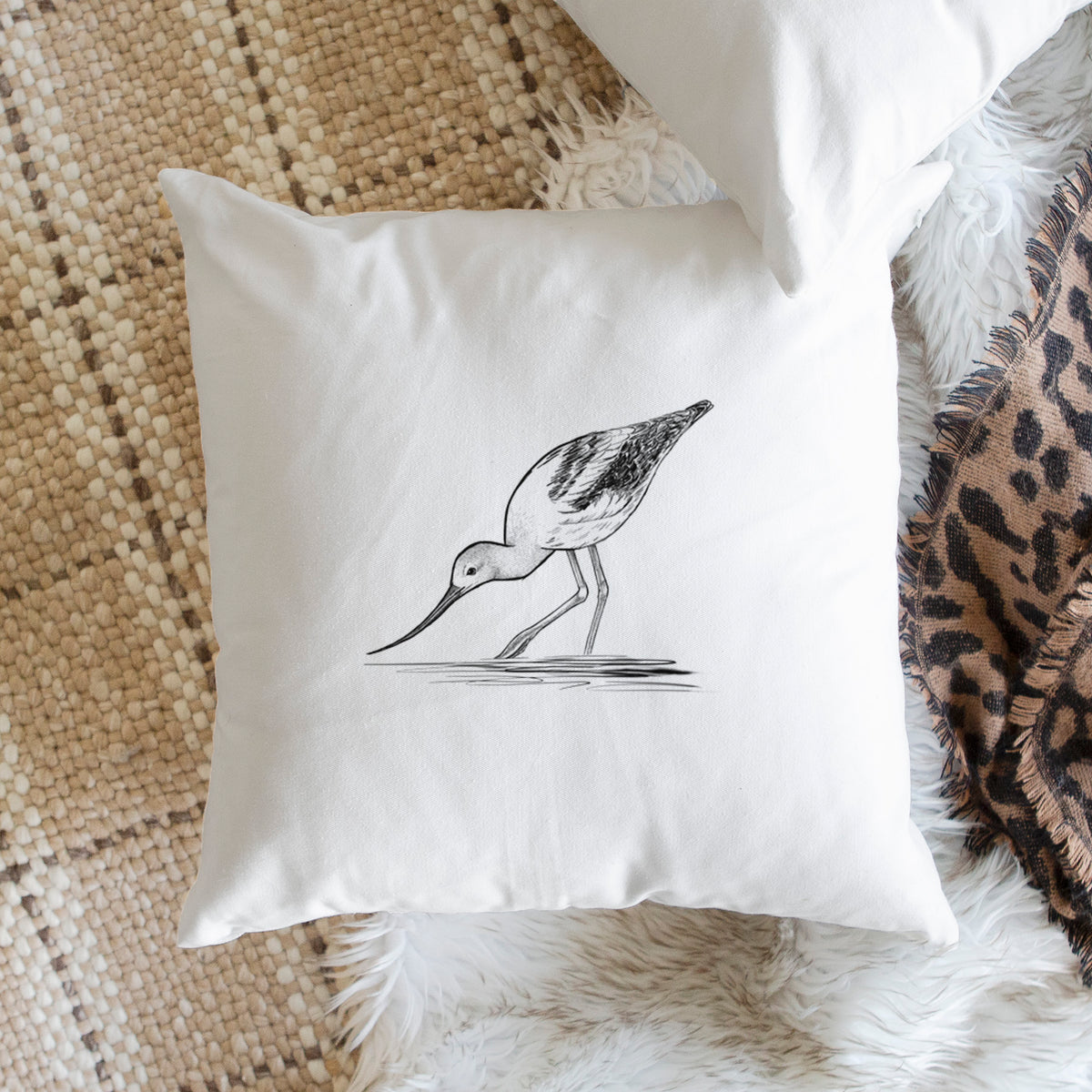 White pillow cover printed with a detailed black ink sketch of an American avocet wading in shallow water, on a woven chair.