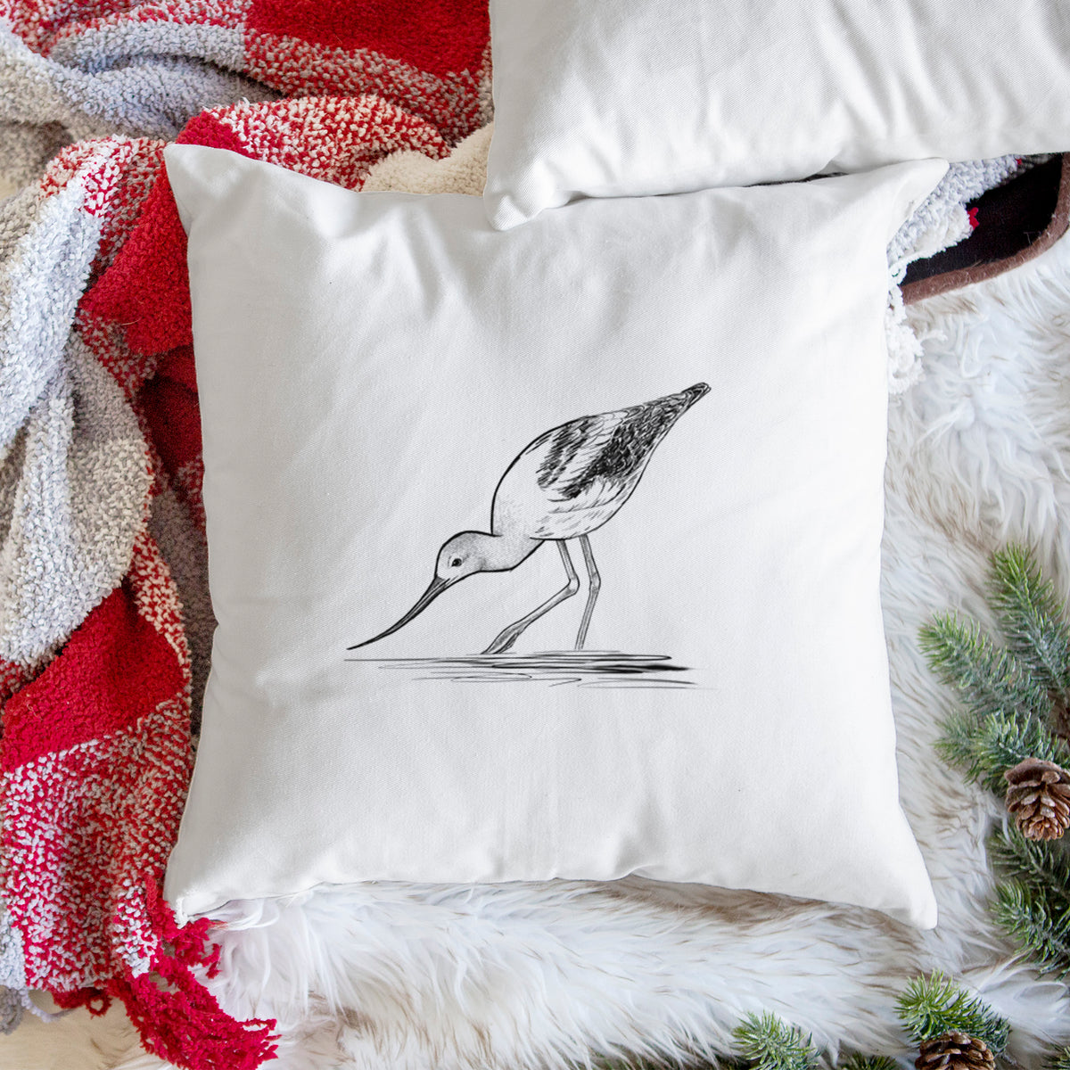 White pillow cover printed with a detailed black ink sketch of an American avocet wading in shallow water, on a woven chair.