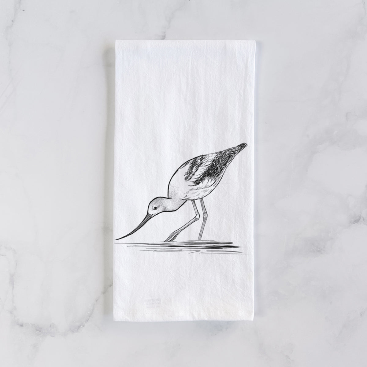White cotton tea towel laid flat on marble, printed with a detailed black ink sketch of an American avocet wading in shallow water.