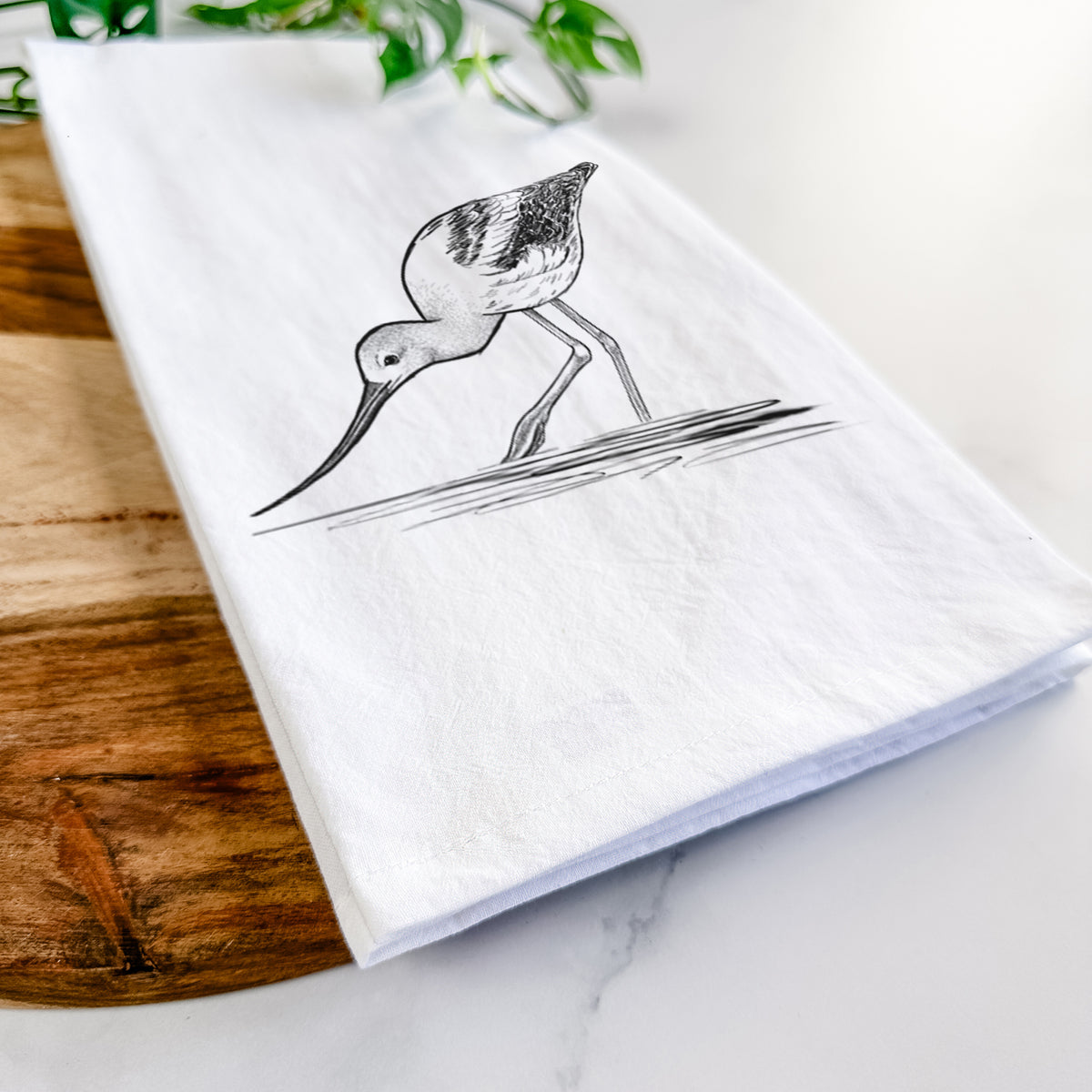Close-up of a folded white tea towel on a wooden board, showing a detailed black ink sketch of an American avocet wading in shallow water.