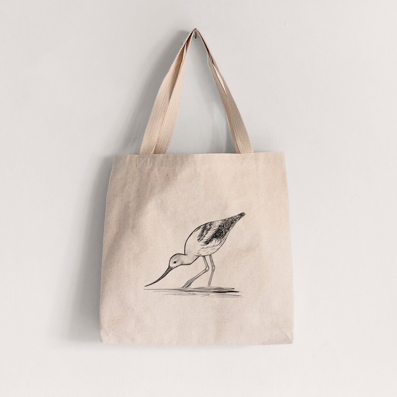Natural canvas tote bag printed with a detailed black ink sketch of an American avocet wading in shallow water.