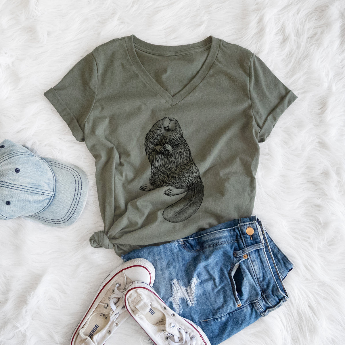 Military green women's relaxed fit cotton V-neck t-shirt printed with a detailed black ink illustration of an American beaver sitting upright with its flat tail, styled flat on a white faux fur rug with a cap, jeans and sneakers.
