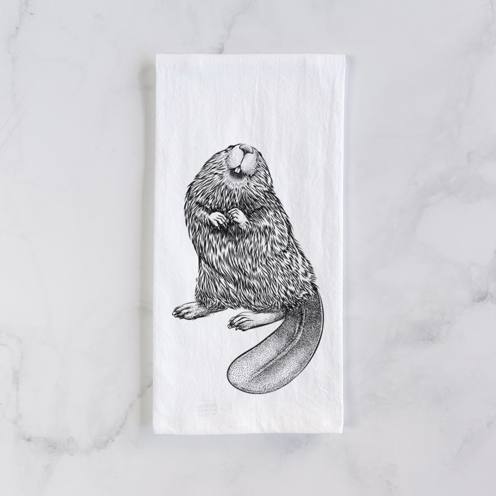 White cotton tea towel printed with a detailed black ink illustration of an American beaver sitting upright with its flat tail, on a marble counter beside a round wooden cutting board and greenery.