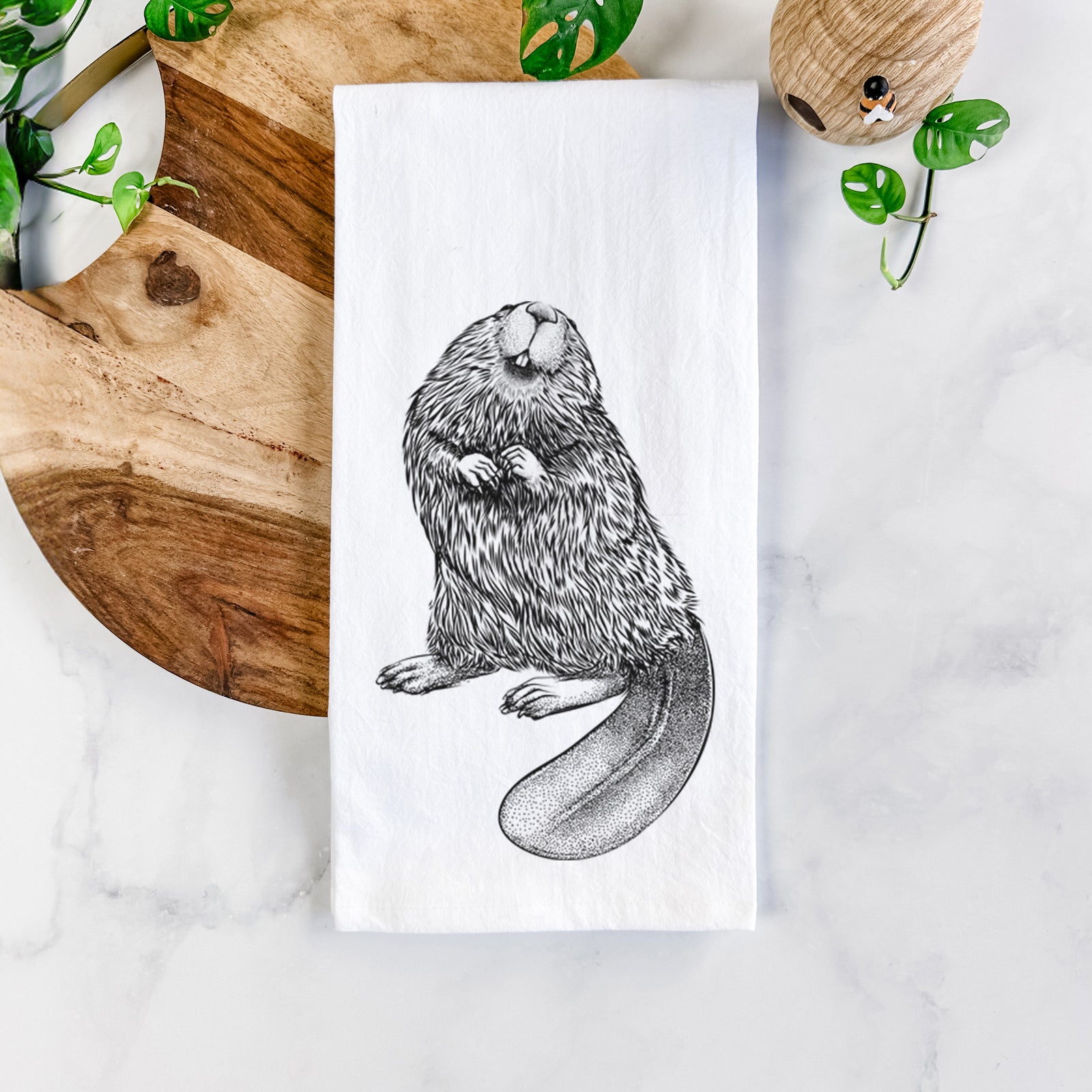 White cotton tea towel printed with a detailed black ink illustration of an American beaver sitting upright with its flat tail, on a marble counter beside a round wooden cutting board and greenery.