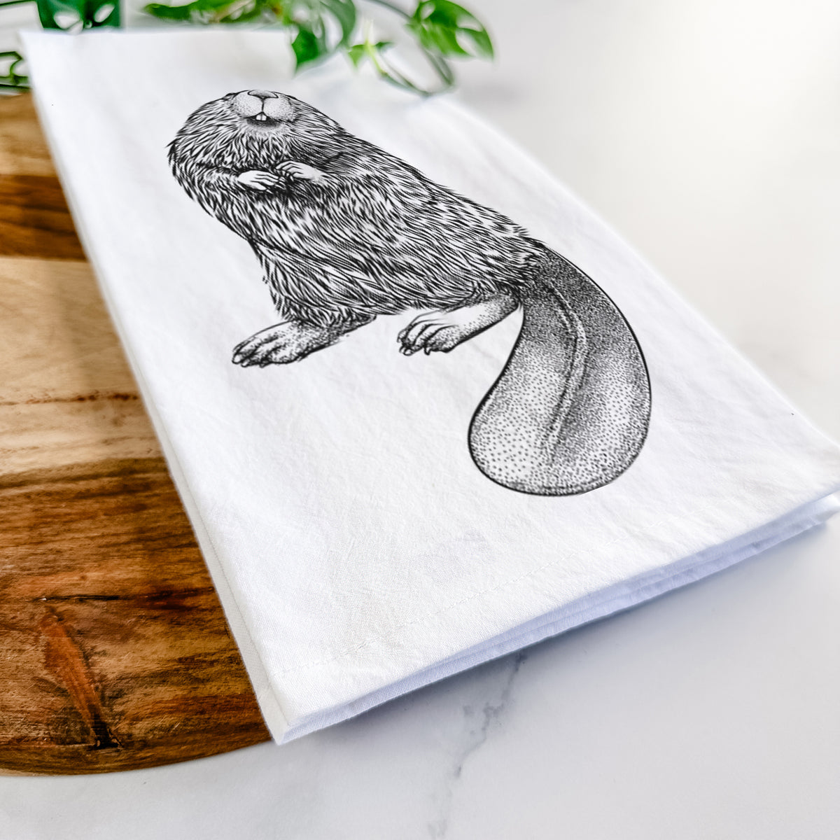 Close-up of a folded white tea towel on a wooden board, showing a detailed black ink illustration of an American beaver sitting upright with its flat tail.