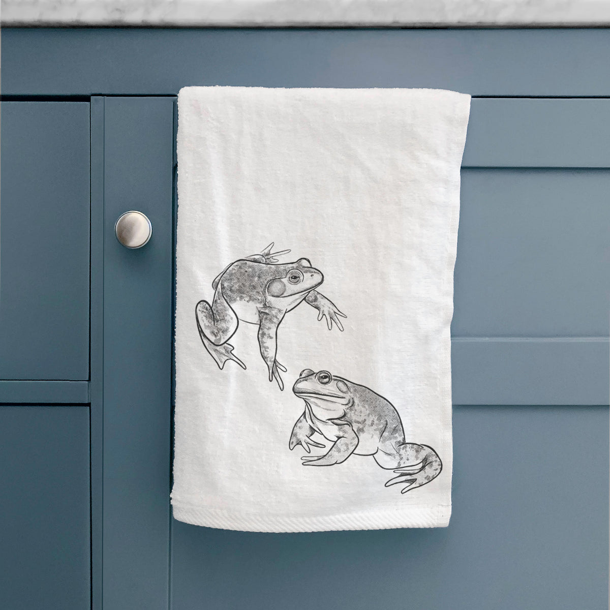 White hand towel printed with a detailed black ink illustration of two American bullfrogs, hanging on a cabinet door.