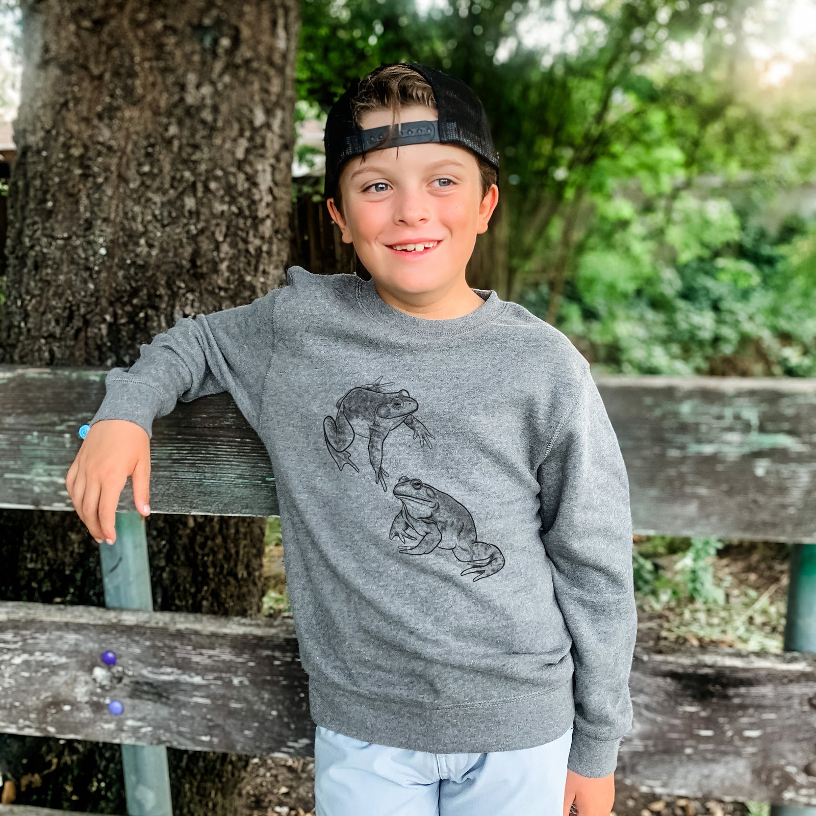 Boy wearing a youth crewneck sweatshirt printed with a detailed black ink illustration of two American bullfrogs, outdoors.