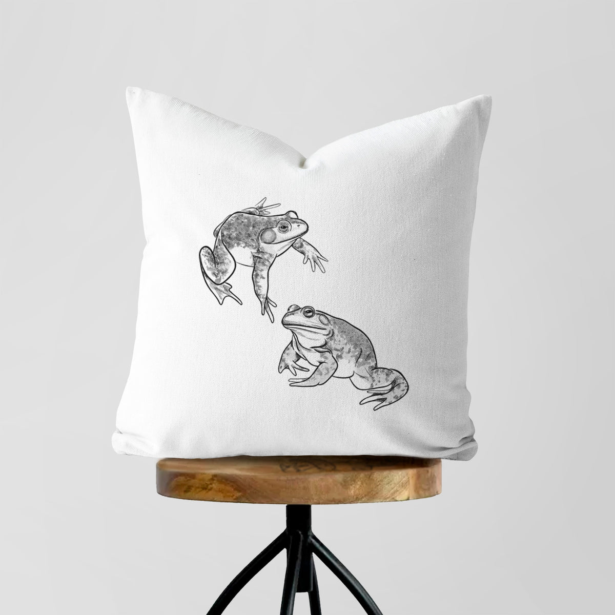 White pillow cover printed with a detailed black ink illustration of two American bullfrogs, on a wooden stool.