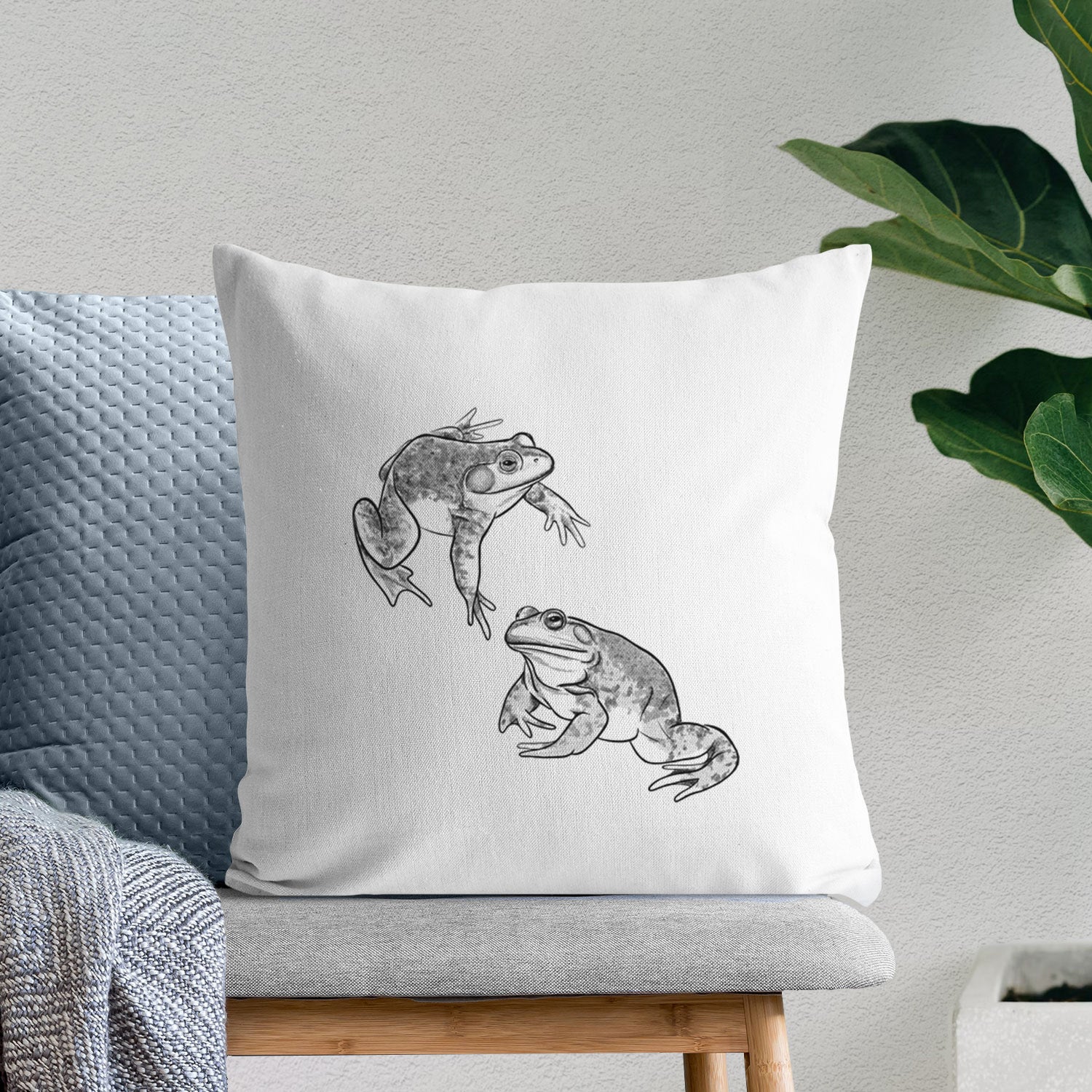 White pillow cover printed with a detailed black ink illustration of two American bullfrogs, on a bench beside plants.