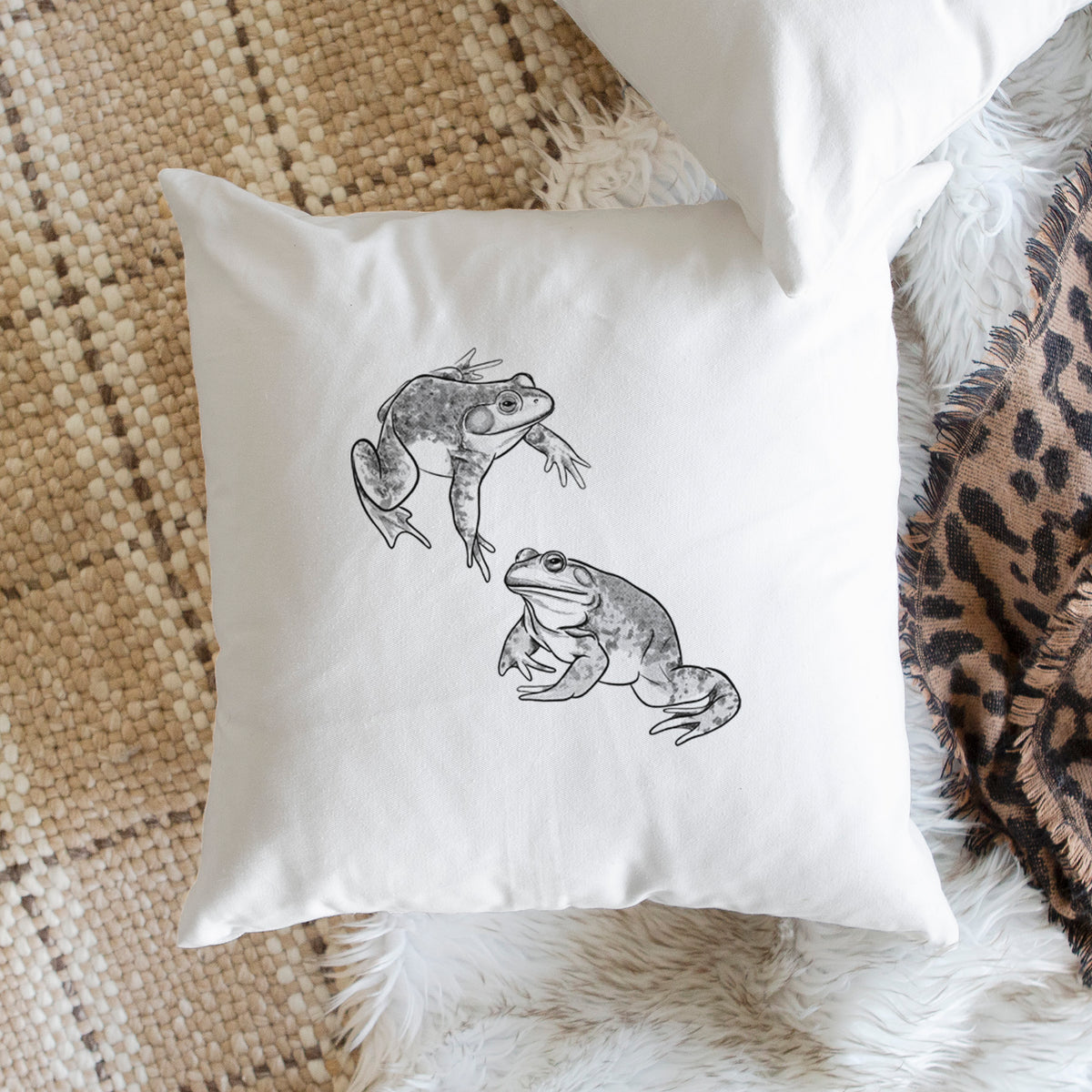 White pillow cover printed with a detailed black ink illustration of two American bullfrogs, on a woven chair.