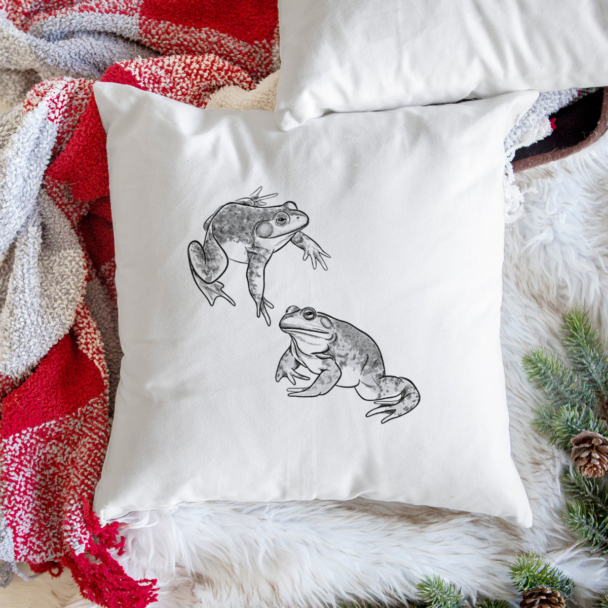 White pillow cover printed with a detailed black ink illustration of two American bullfrogs, on a woven chair.