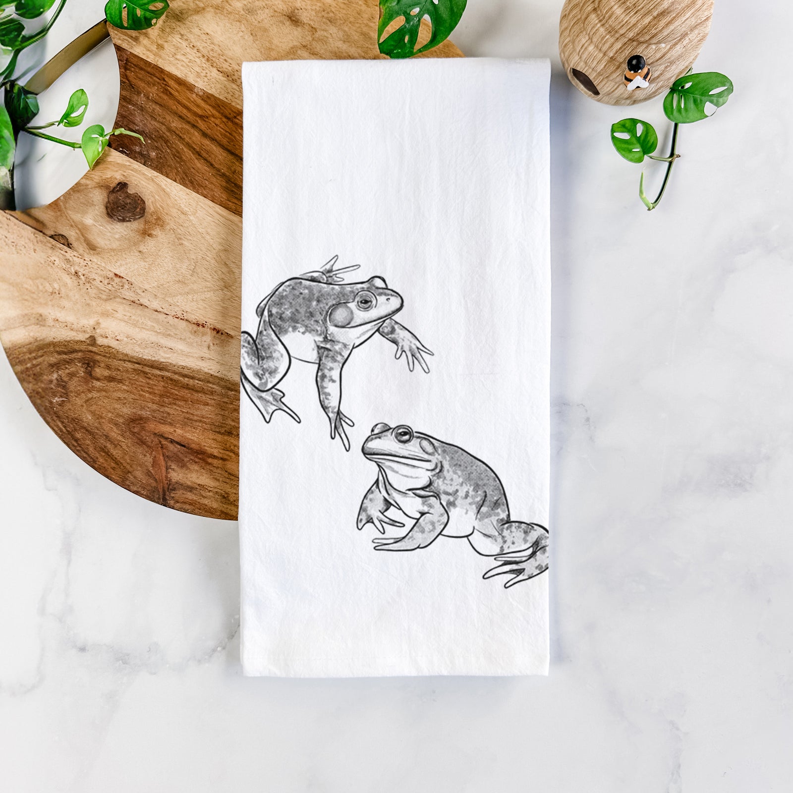 White cotton tea towel printed with a detailed black ink illustration of two American bullfrogs, on a marble counter beside a round wooden cutting board and greenery.