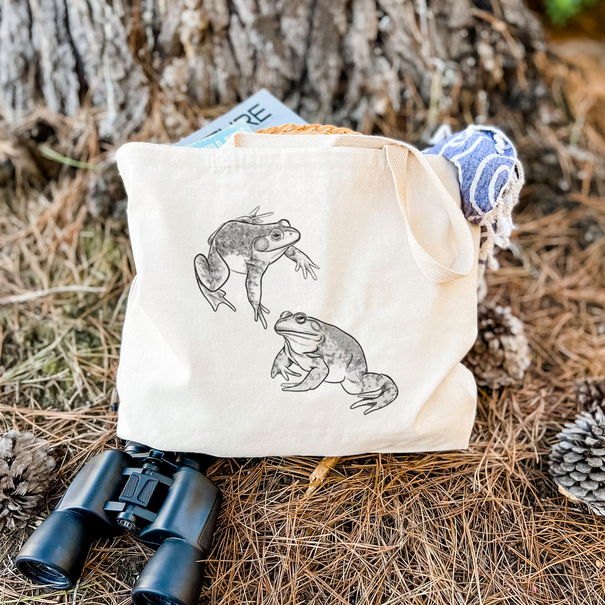 Canvas tote bag printed with a detailed black ink illustration of two American bullfrogs, resting on dry grass beside a pair of binoculars.