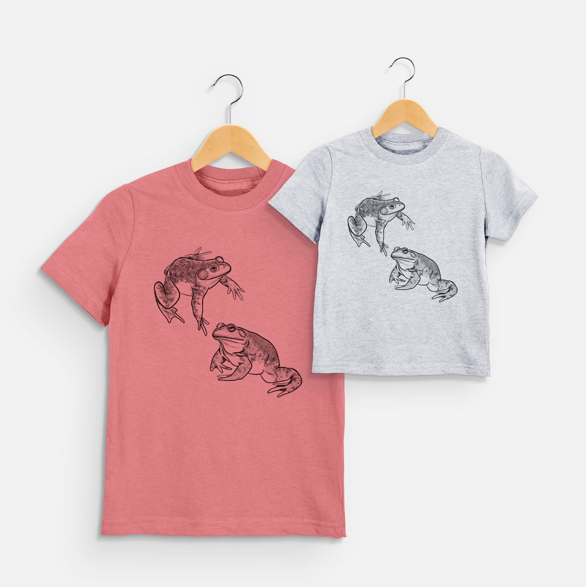 Two youth t-shirts in different colors on hangers, printed with a detailed black ink illustration of two American bullfrogs.