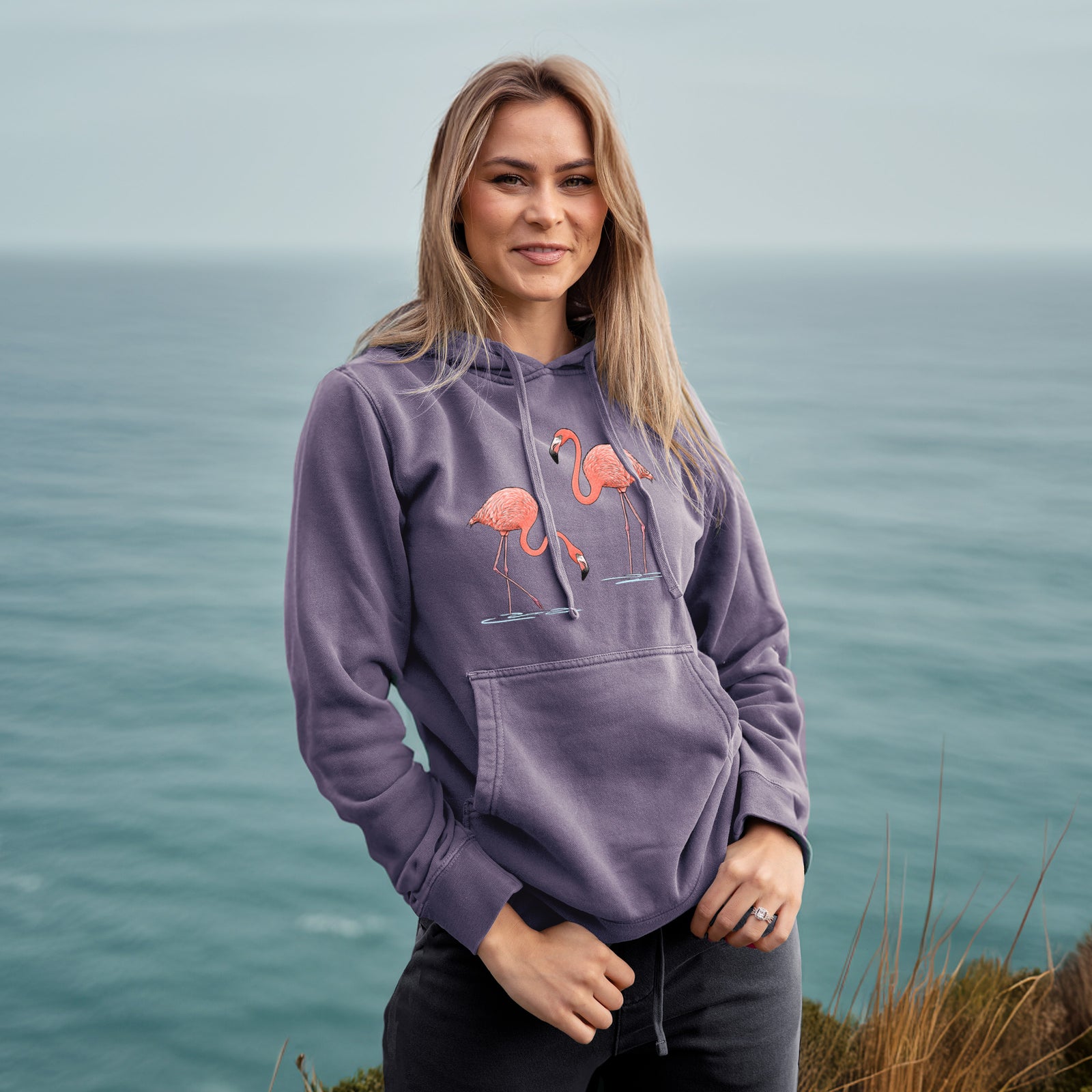 Plum purple pigment dyed hoodie laid flat, printed with two pink American flamingos wading in water, one feeding with its head down and one standing tall.