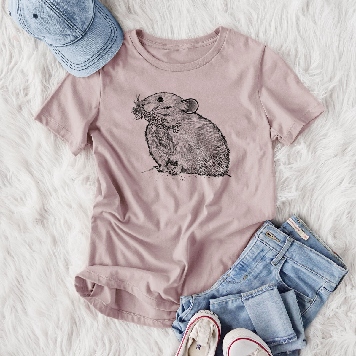 Women's relaxed fit cotton t-shirt printed with a detailed black ink illustration of an American pika holding flowers and grass in its mouth, styled flat on a white faux fur rug with jeans.