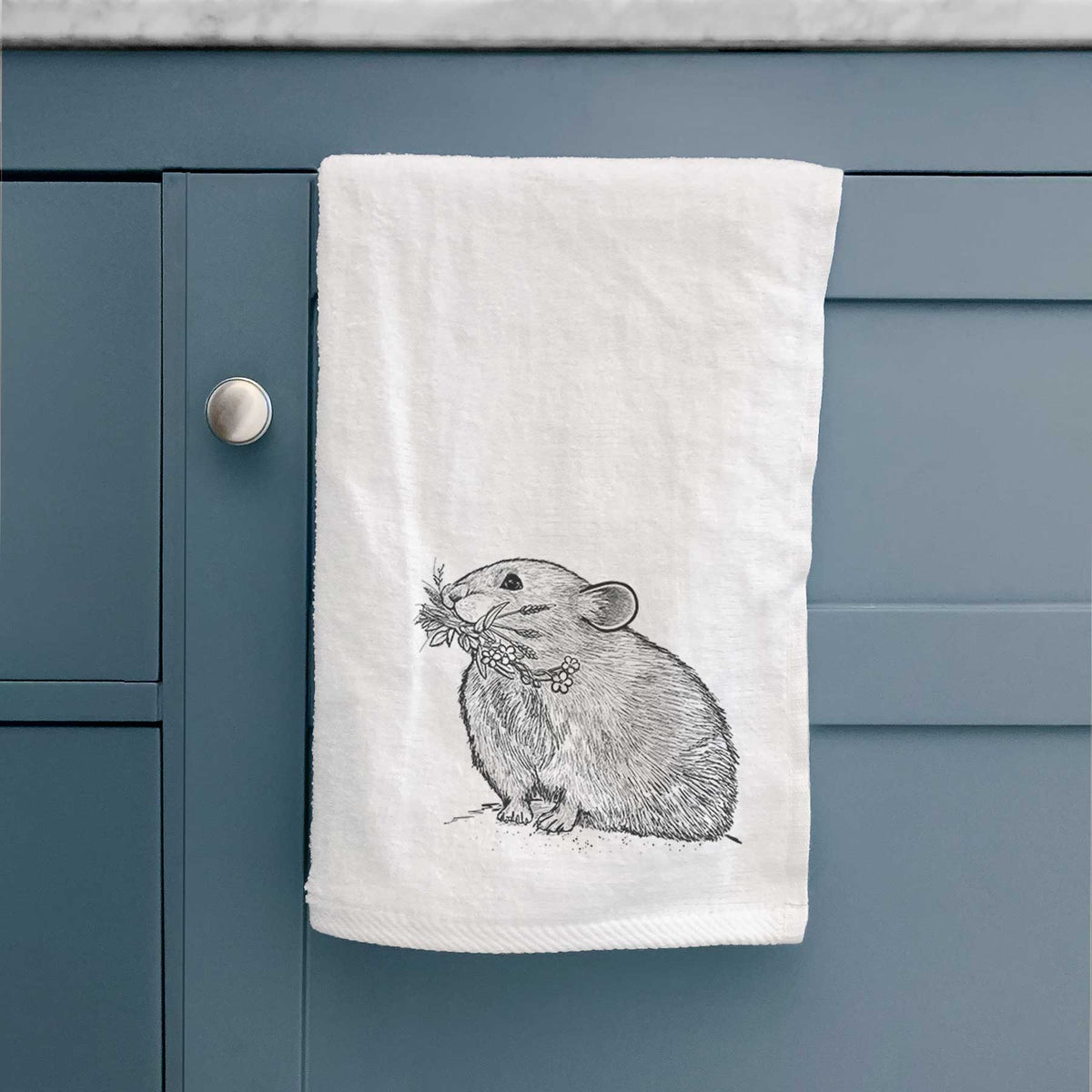 White hand towel printed with a detailed black ink illustration of an American pika holding flowers and grass in its mouth, hanging on a cabinet door.