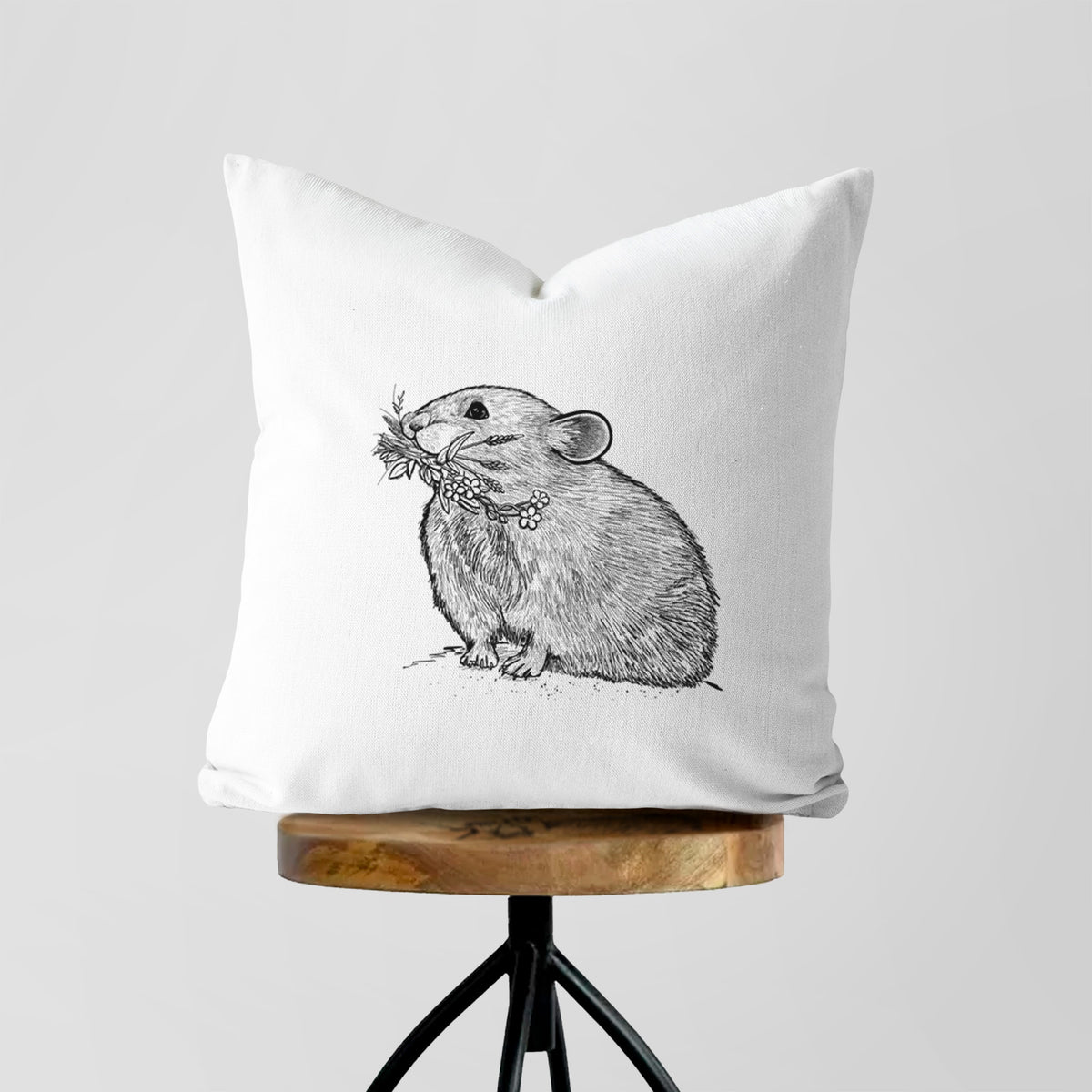 White pillow cover printed with a detailed black ink illustration of an American pika holding flowers and grass in its mouth, on a wooden stool.