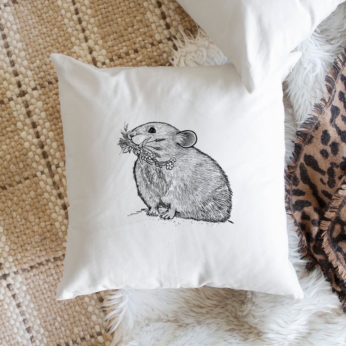 White pillow cover printed with a detailed black ink illustration of an American pika holding flowers and grass in its mouth, on a woven chair.