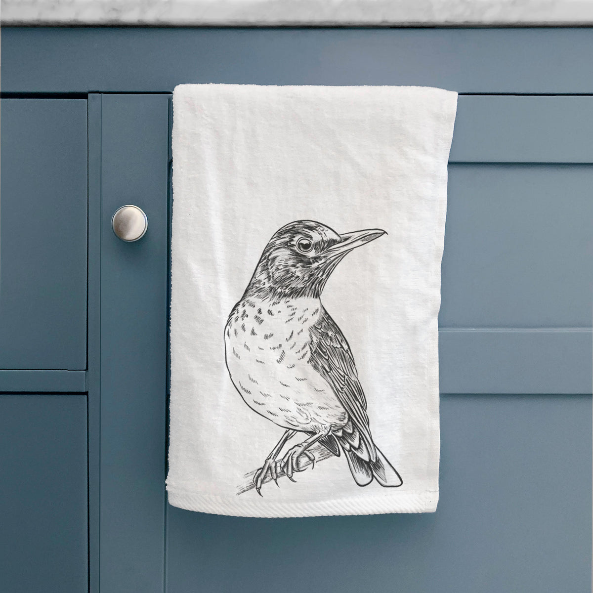 White hand towel printed with a detailed black ink sketch of an American robin perched on a branch, hanging on a cabinet door.