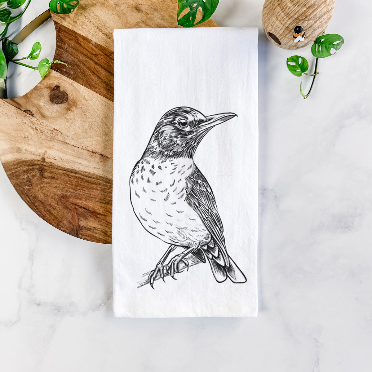 White cotton tea towel printed with a detailed black ink sketch of an American robin perched on a branch, on a marble counter beside a round wooden cutting board and greenery.