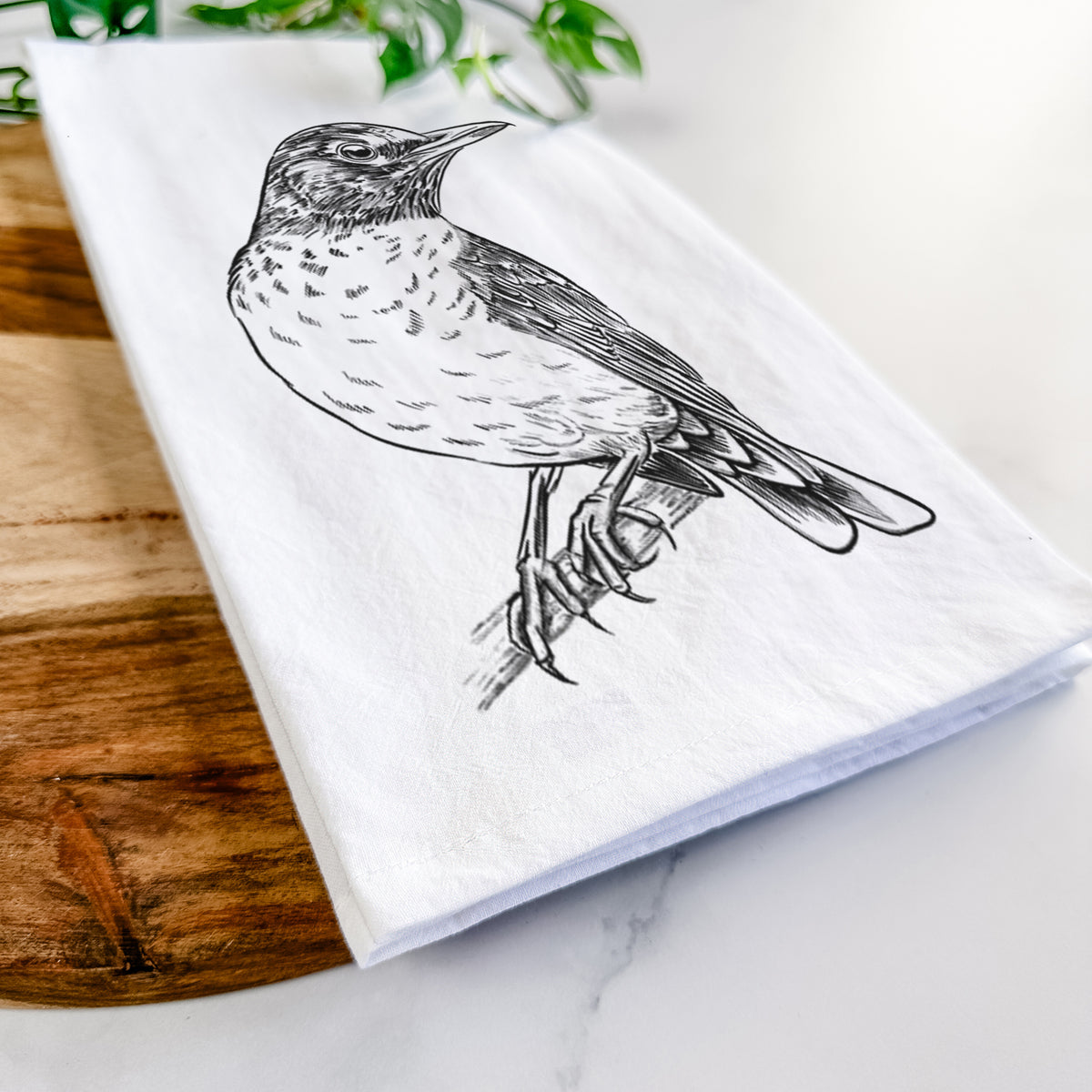 Close-up of a folded white tea towel on a wooden board, showing a detailed black ink sketch of an American robin perched on a branch.