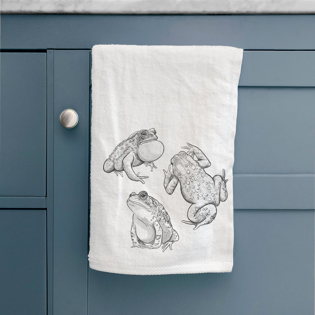 White hand towel printed with a black ink illustration of three American toads, hanging on a cabinet door.
