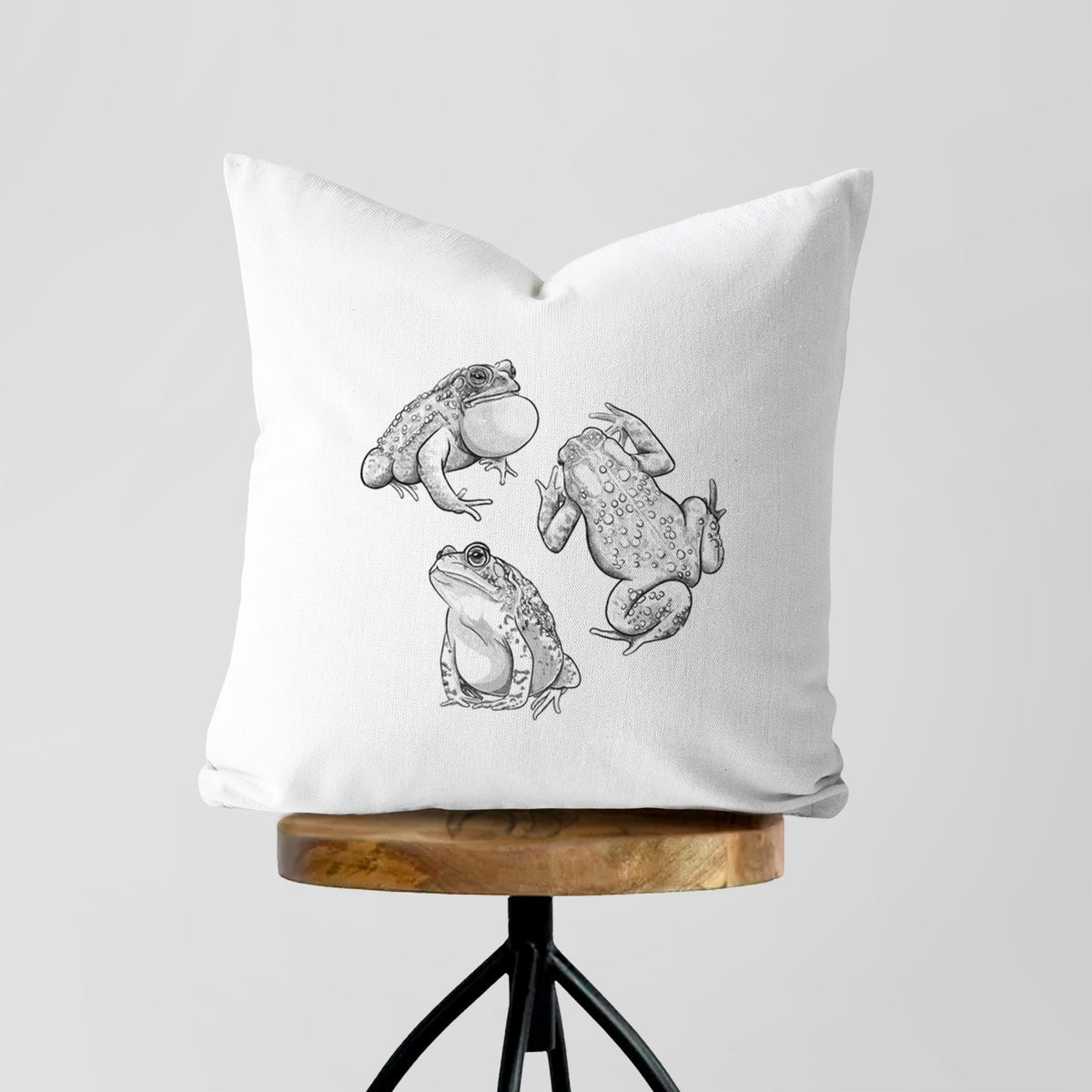 White pillow cover printed with a black ink illustration of three American toads, on a wooden stool.
