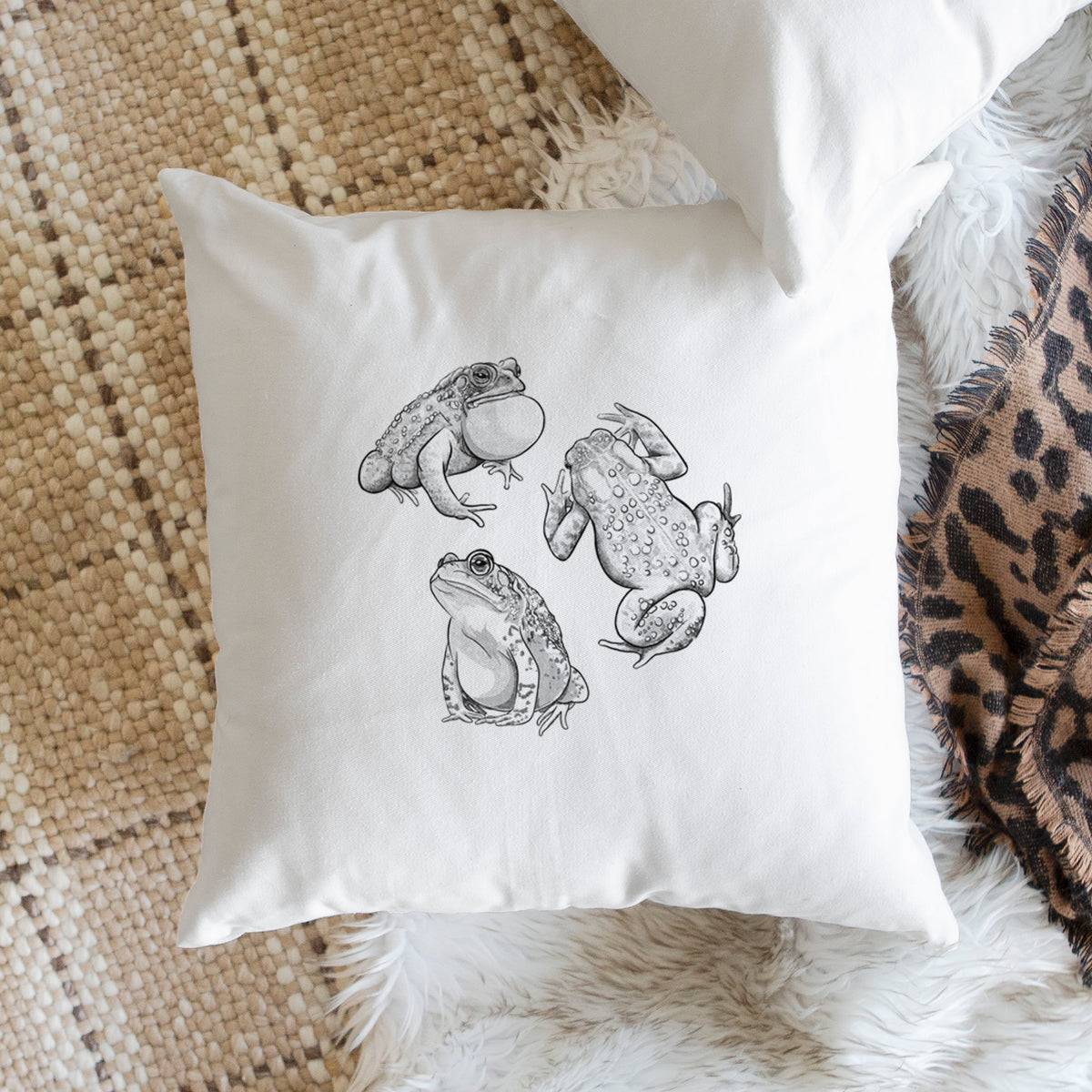 White pillow cover printed with a black ink illustration of three American toads, on a woven chair.