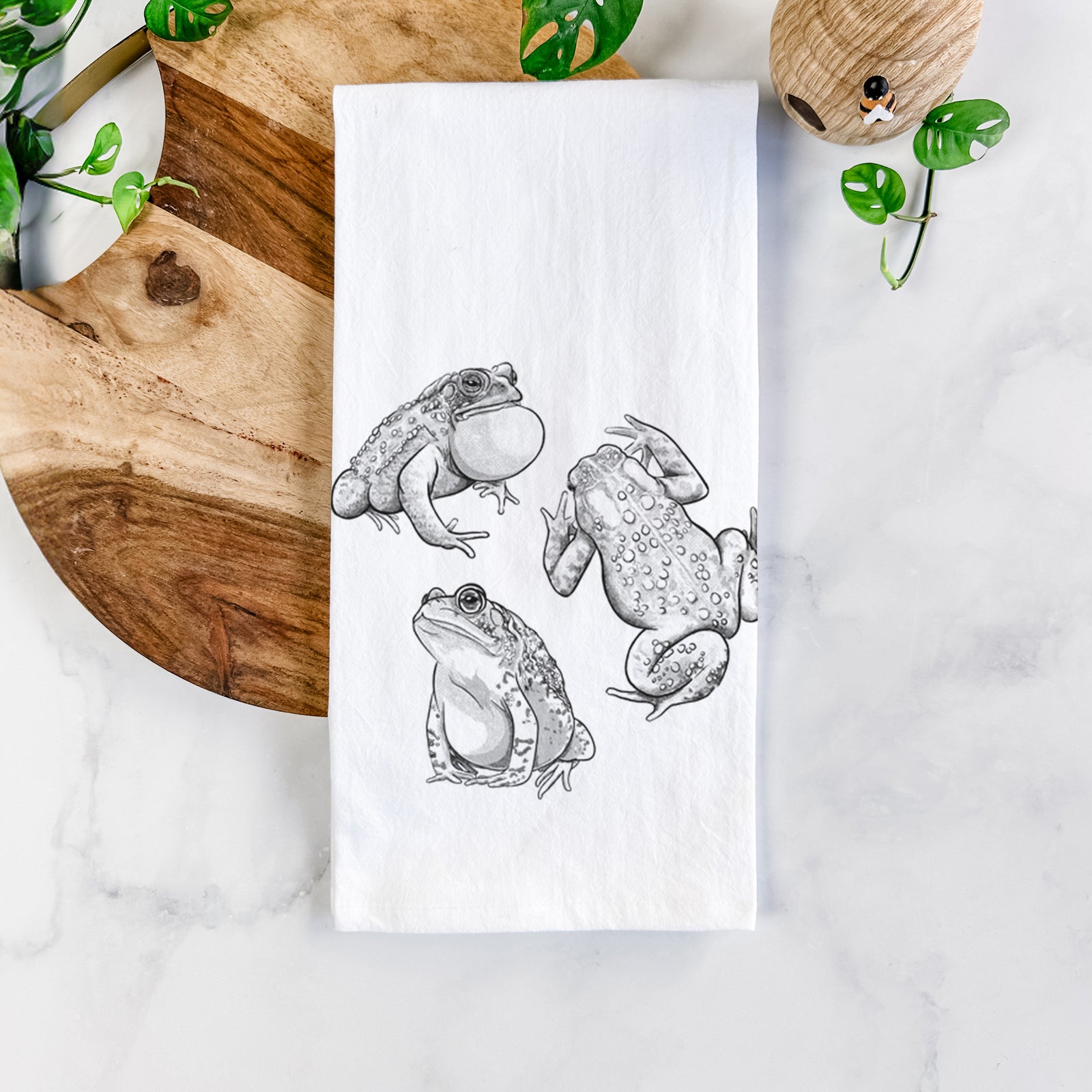 White cotton tea towel printed with a black ink illustration of three American toads, on a marble counter beside a round wooden cutting board and greenery.
