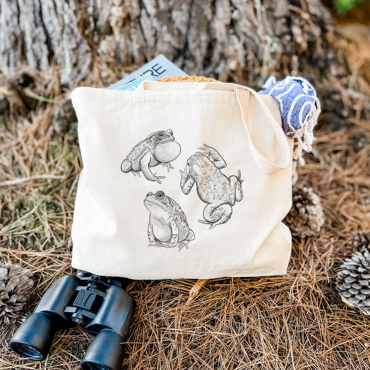 Canvas tote bag printed with a black ink illustration of three American toads, resting on dry grass beside a pair of binoculars.