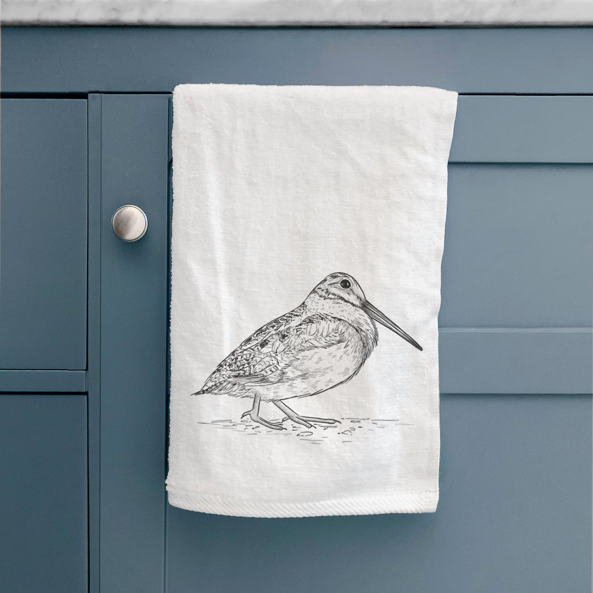 White hand towel printed with a detailed black ink sketch of an American woodcock standing on the ground, hanging on a cabinet door.