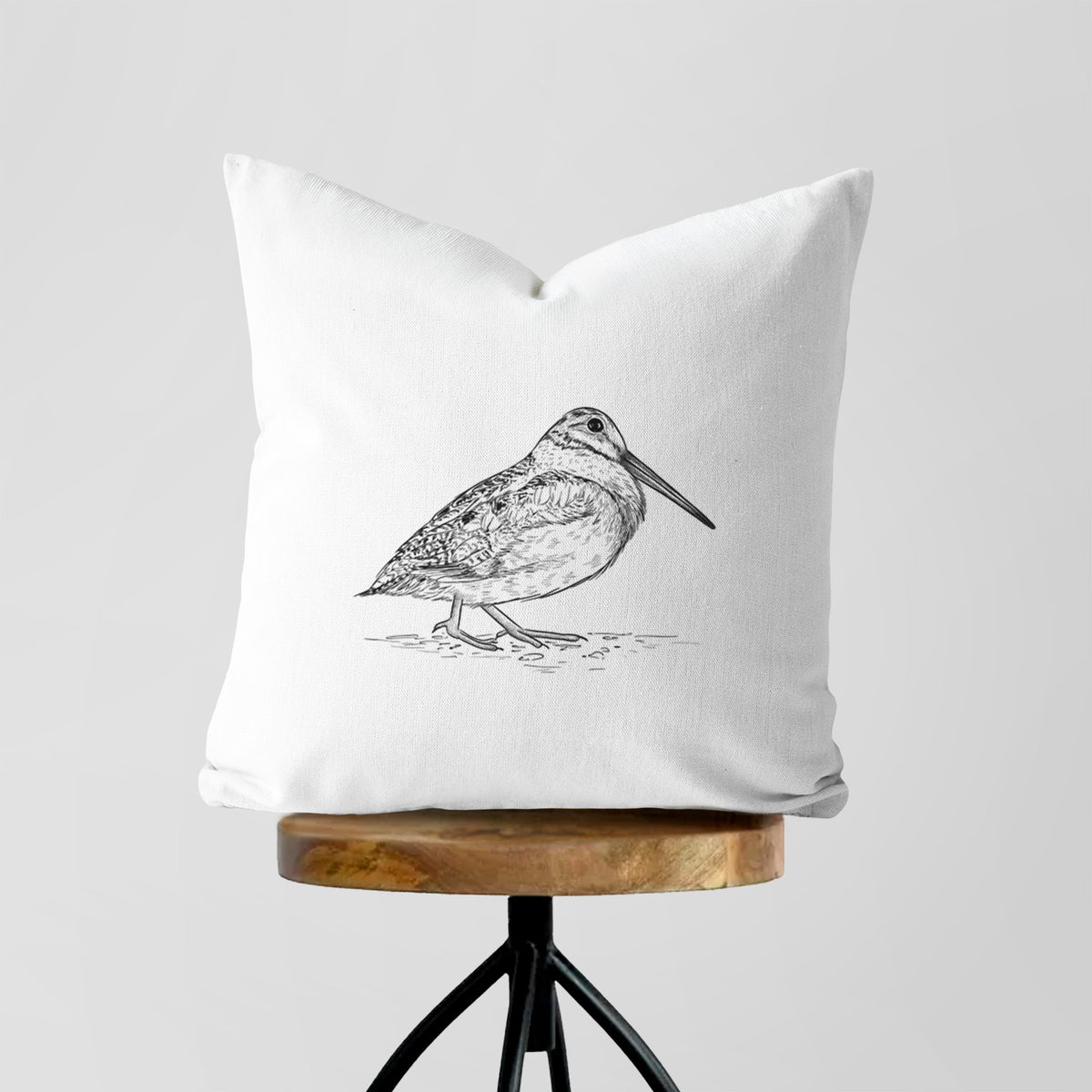 White pillow cover printed with a detailed black ink sketch of an American woodcock standing on the ground, on a wooden stool.