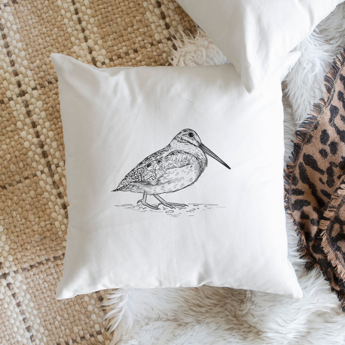 White pillow cover printed with a detailed black ink sketch of an American woodcock standing on the ground, on a woven chair.