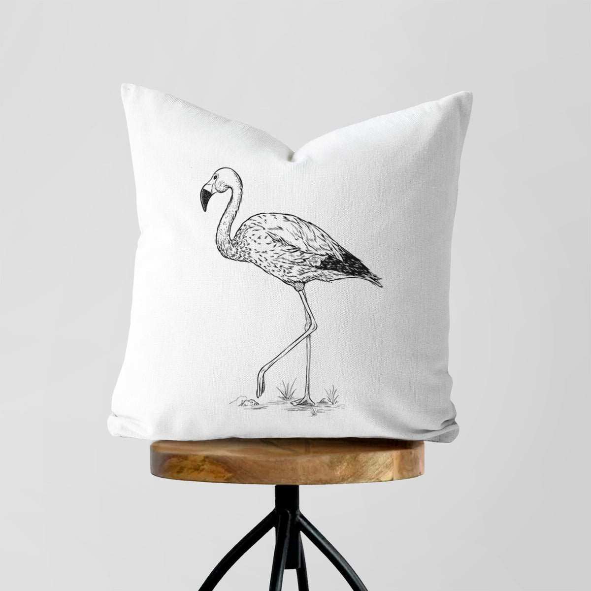 White pillow cover printed with a black ink illustration of an Andean flamingo standing on one leg, on a wooden stool.