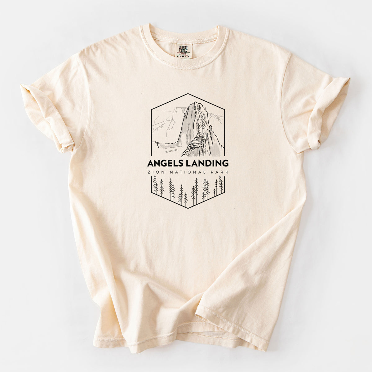 Ivory Comfort Colors t-shirt laid flat, printed with a black line art badge of Angels Landing above pine trees, with the words Angels Landing, Zion National Park.