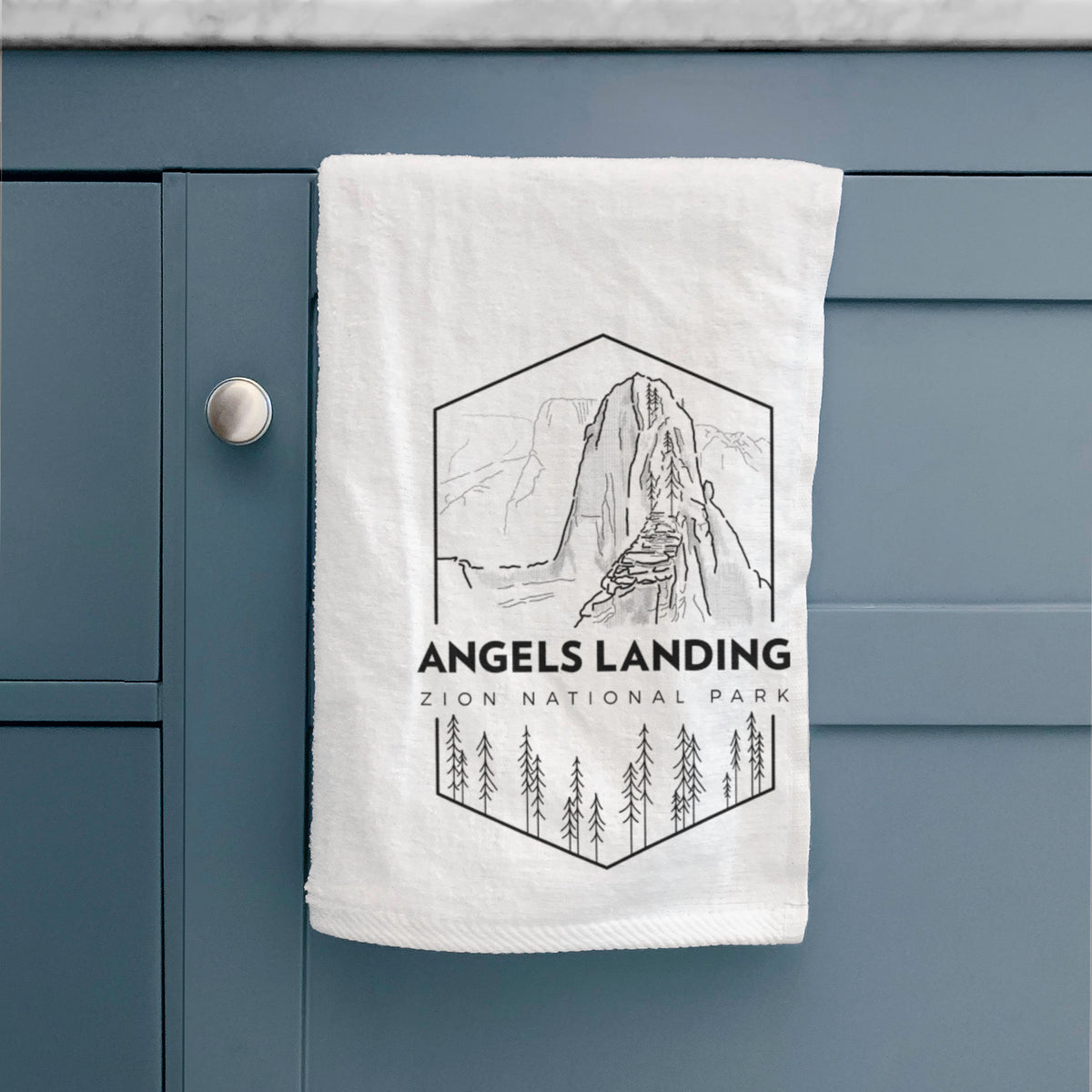 White hand towel printed with a black line art badge of Angels Landing above pine trees, with the words Angels Landing, Zion National Park, hanging on a cabinet door.